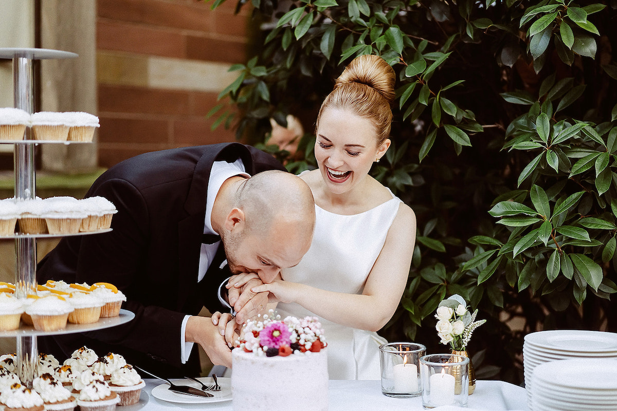 Bräutigam beißt in die Hand seiner Braut als diese versucht die Hochzeitstorte anzuschneiden