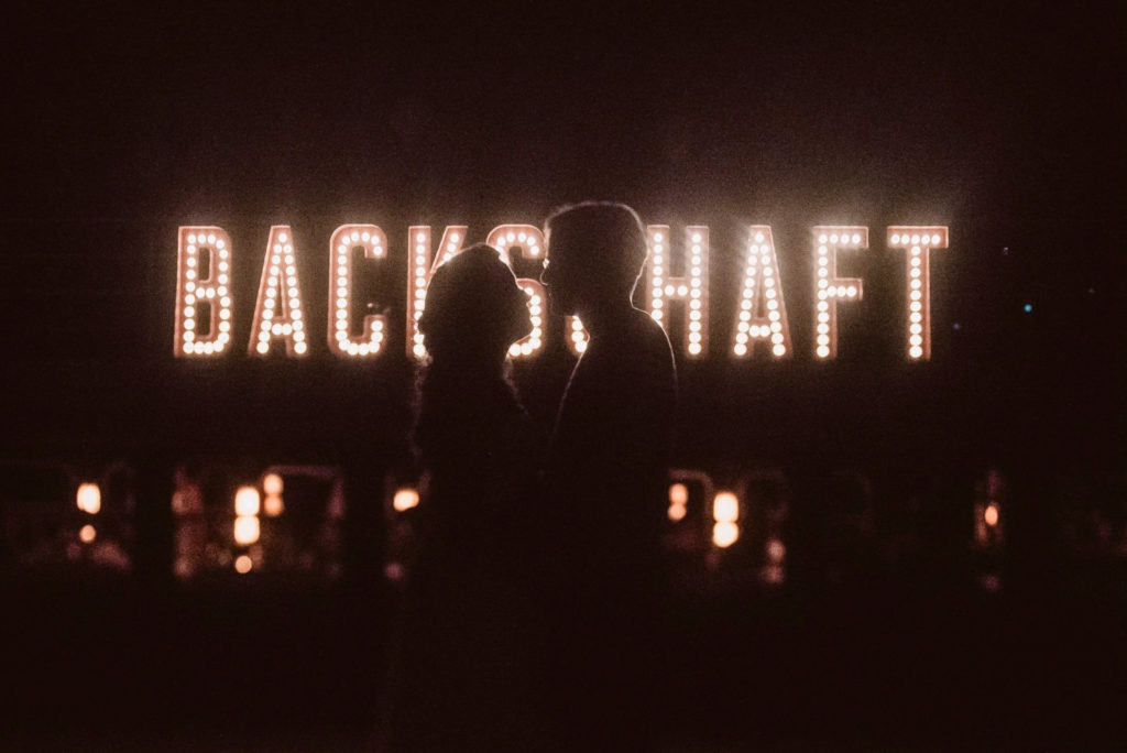 Das Brautpaar steht in Silhouette vor dem leuchtenden Backschaft-Schriftzug zwischen Kerzen.
