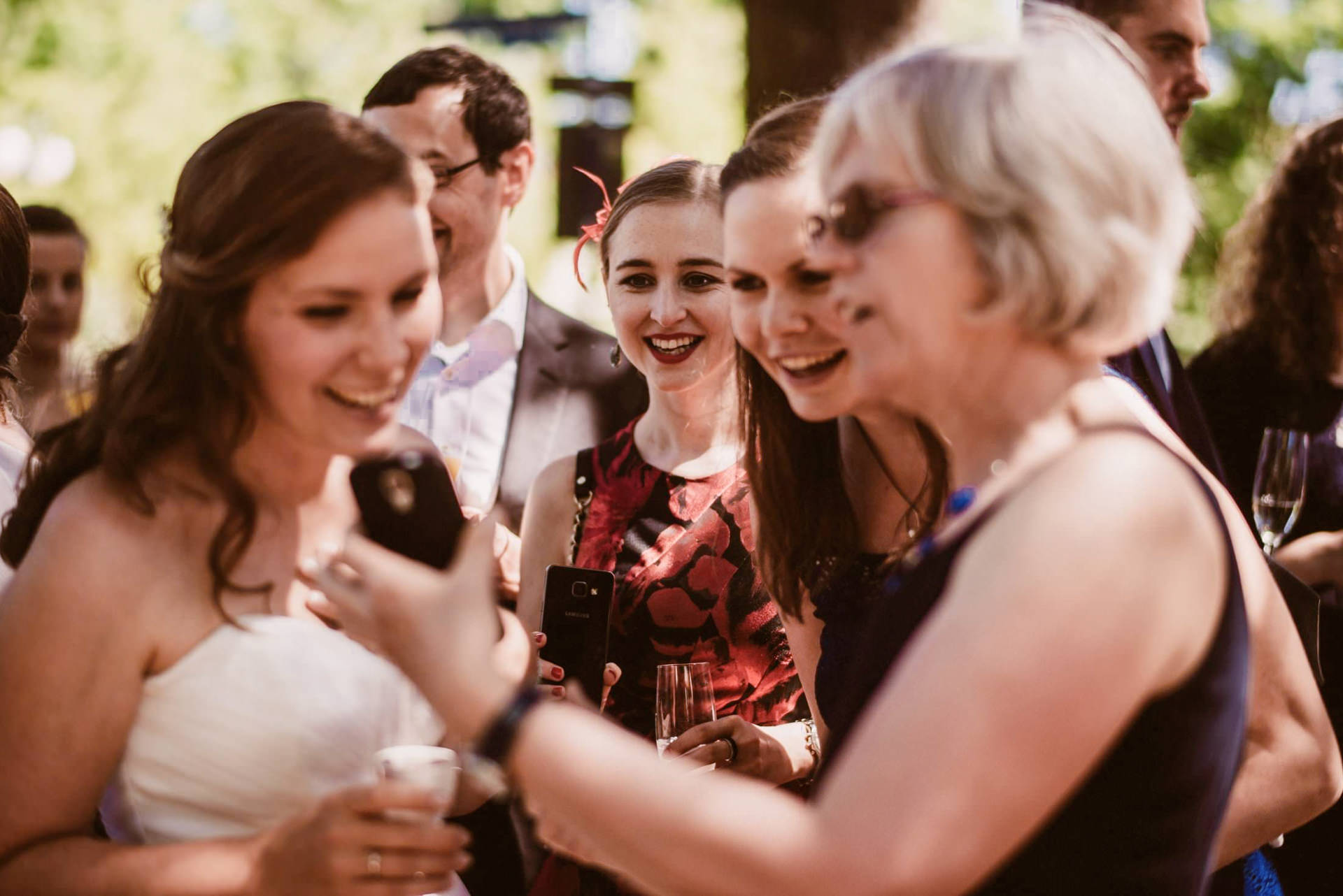 Hochzeitsgäste lachen und stoßen an mit Champagner, Konfetti und Luftballons unter Bäumen.