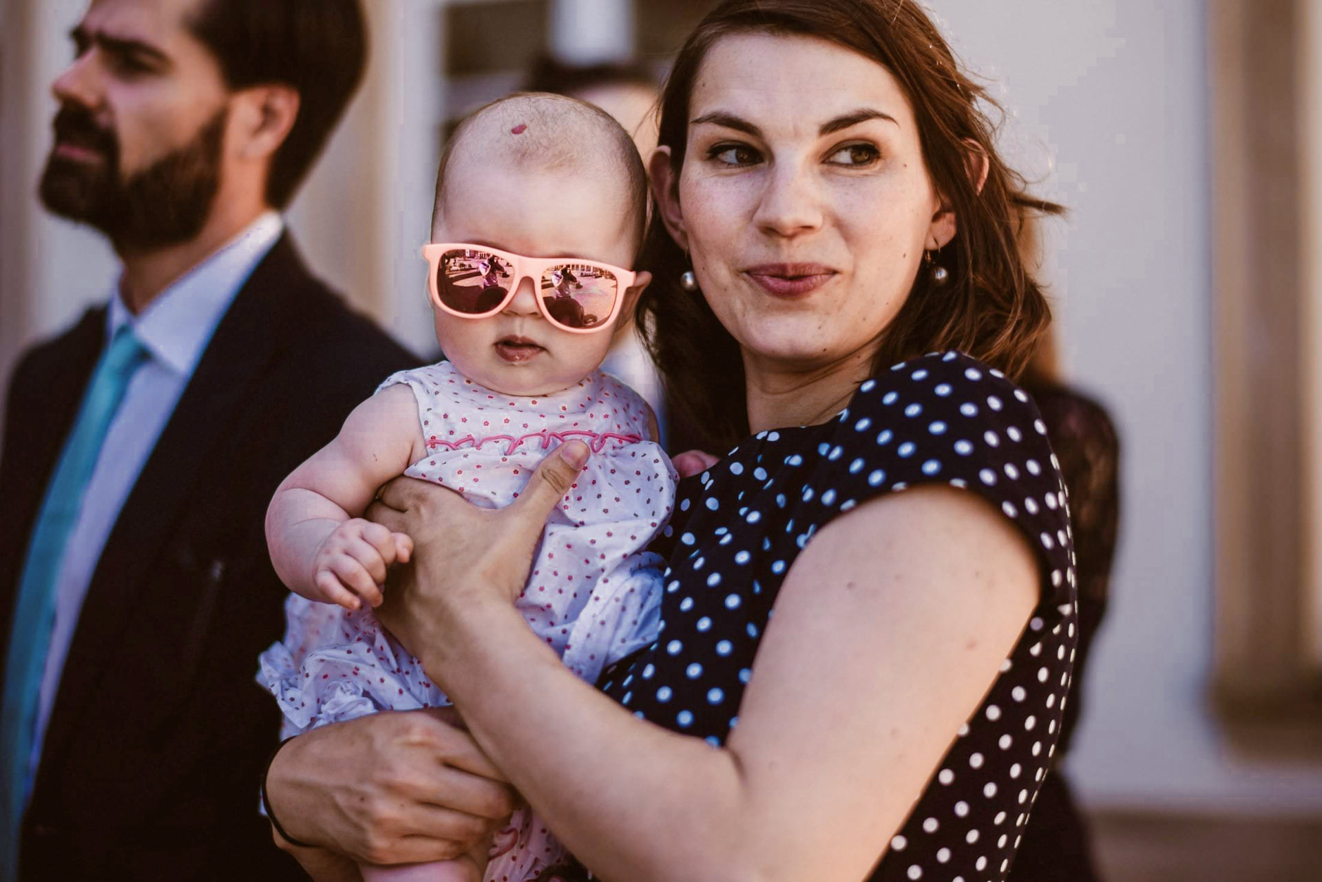 Eine Frau in gepunkteter Bluse hält ein lachendes, überraschtes Baby mit lustiger Sonnenbrille.