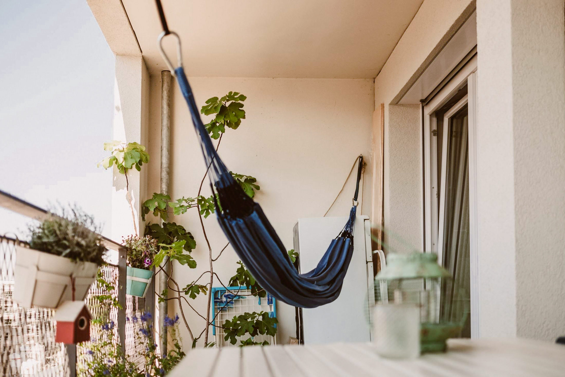 Eine Hängematte hängt auf einem hellen Balkon umgeben von grünem Laub.