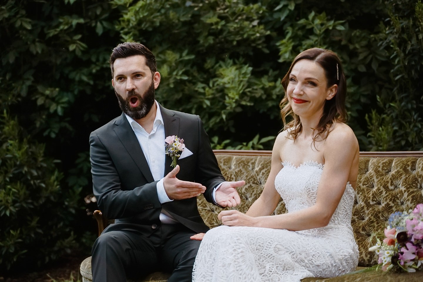 Brautpaar auf einem Vintage-Sofa im Grünen, der Bräutigam erzählt gestikulierend, die Braut lacht – Hochzeit im Berthold 57 Kraichtal
