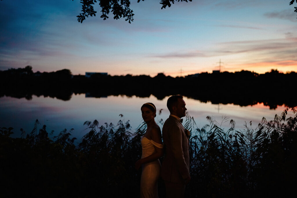 Brautpaar steht als Silhouette am Seeufer im Abendrot, das Wasser spiegelt den Himmel.