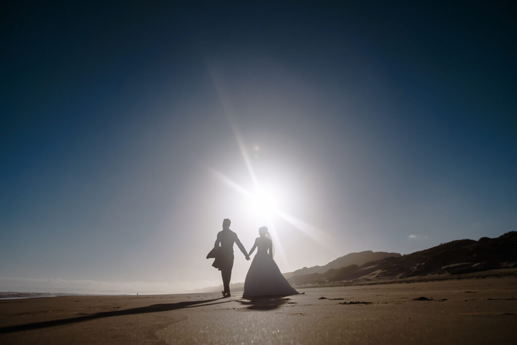Brautpaar geht Hand in Hand am Strand, Sonne blendet hell im Hintergrund