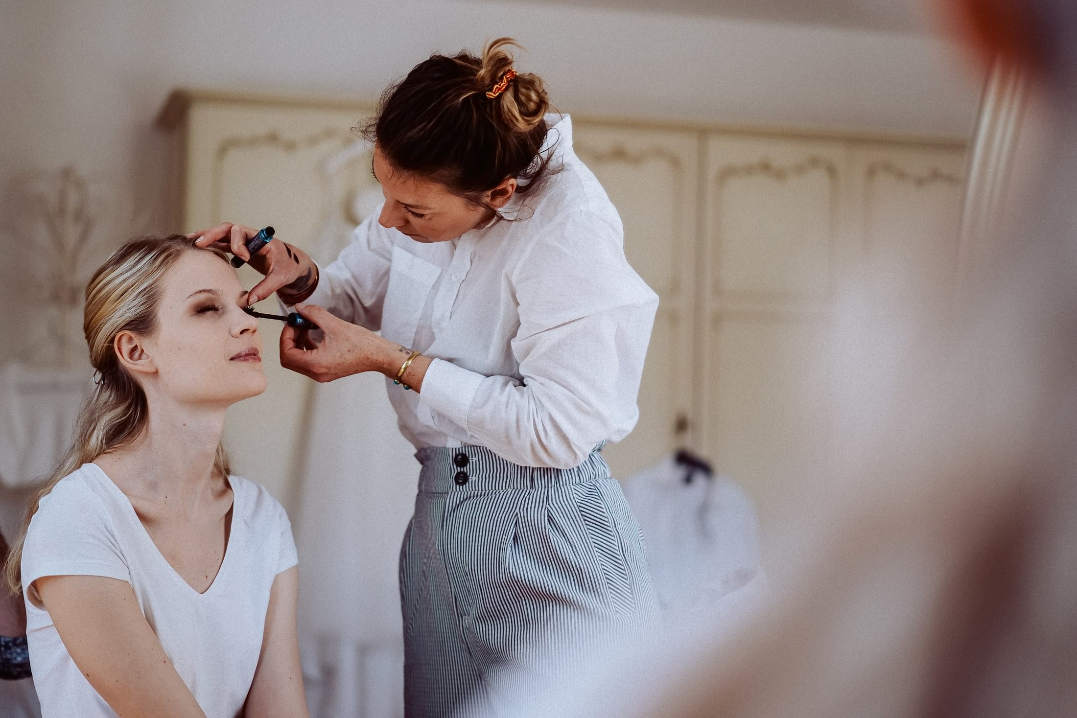 Einer sitzenden Frau wird mit einem Wimpernstäbchen Make-up am Auge aufgetragen.