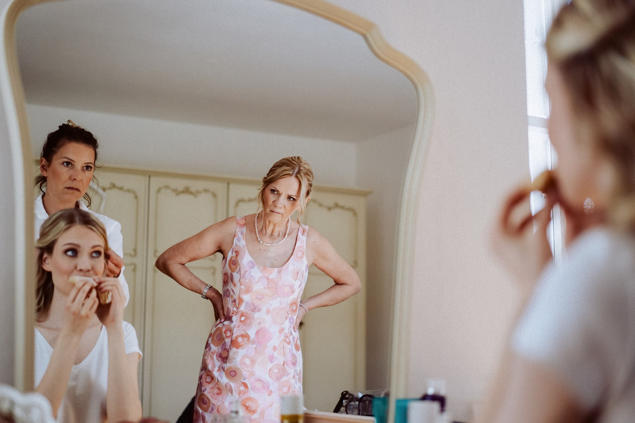Eine Frau im geblümten Kleid prüft ihr Spiegelbild, im Hintergrund zwei junge Frauen.
