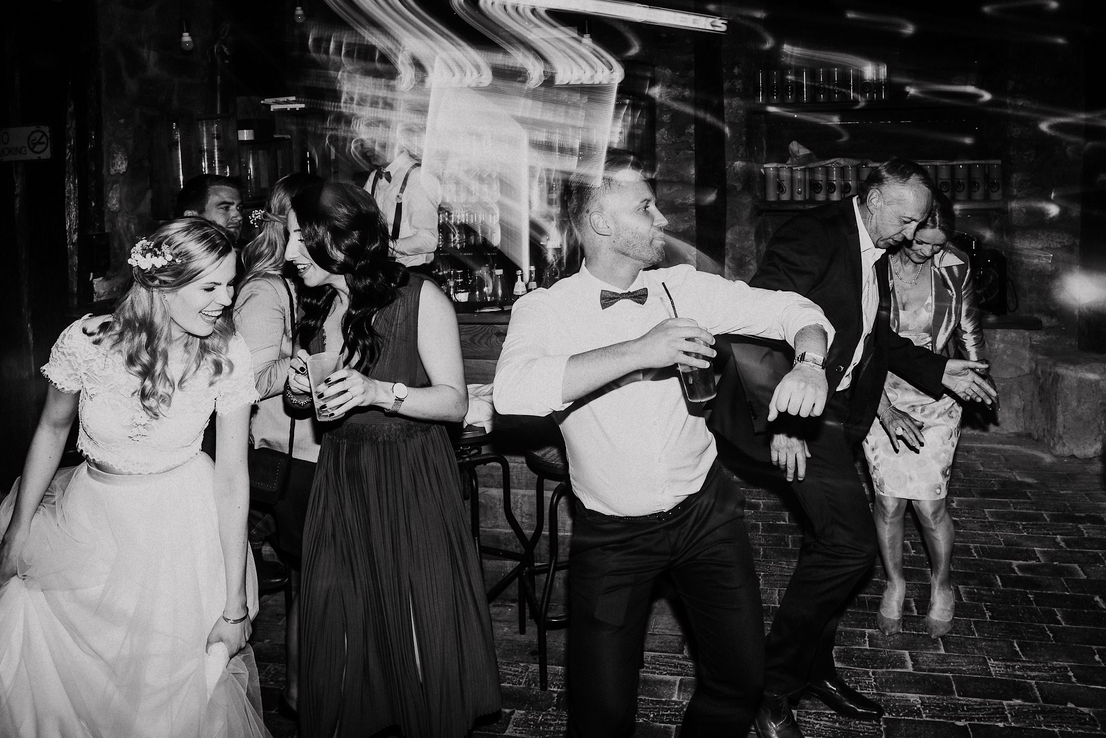 An der Bar lacht die Braut mit einer Gästin, während ein Mann tanzend vorbeizieht.