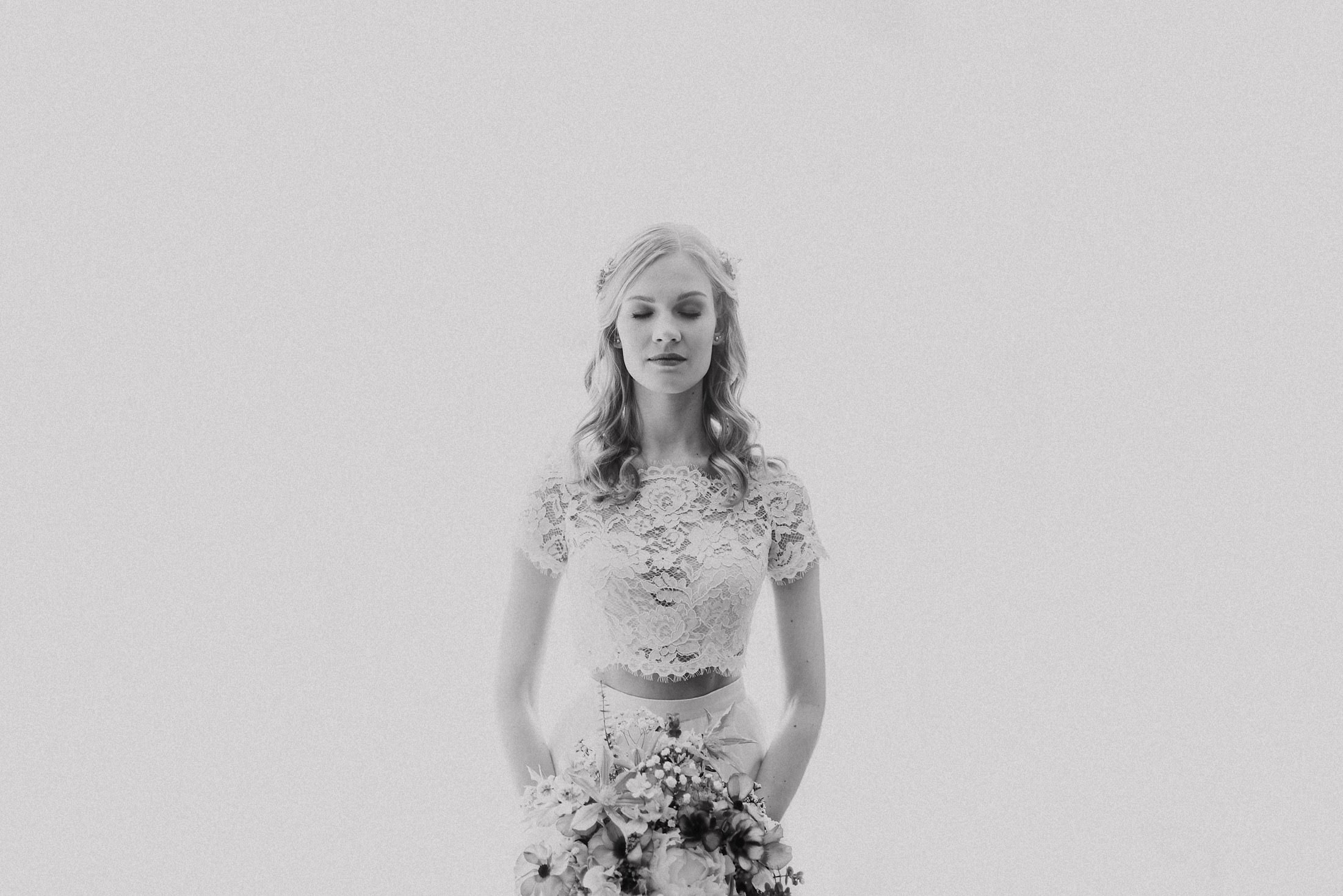Die Braut im Spitzen-Crop-Top mit Blumen im Haar und Brautstrauß, Augen geschlossen.