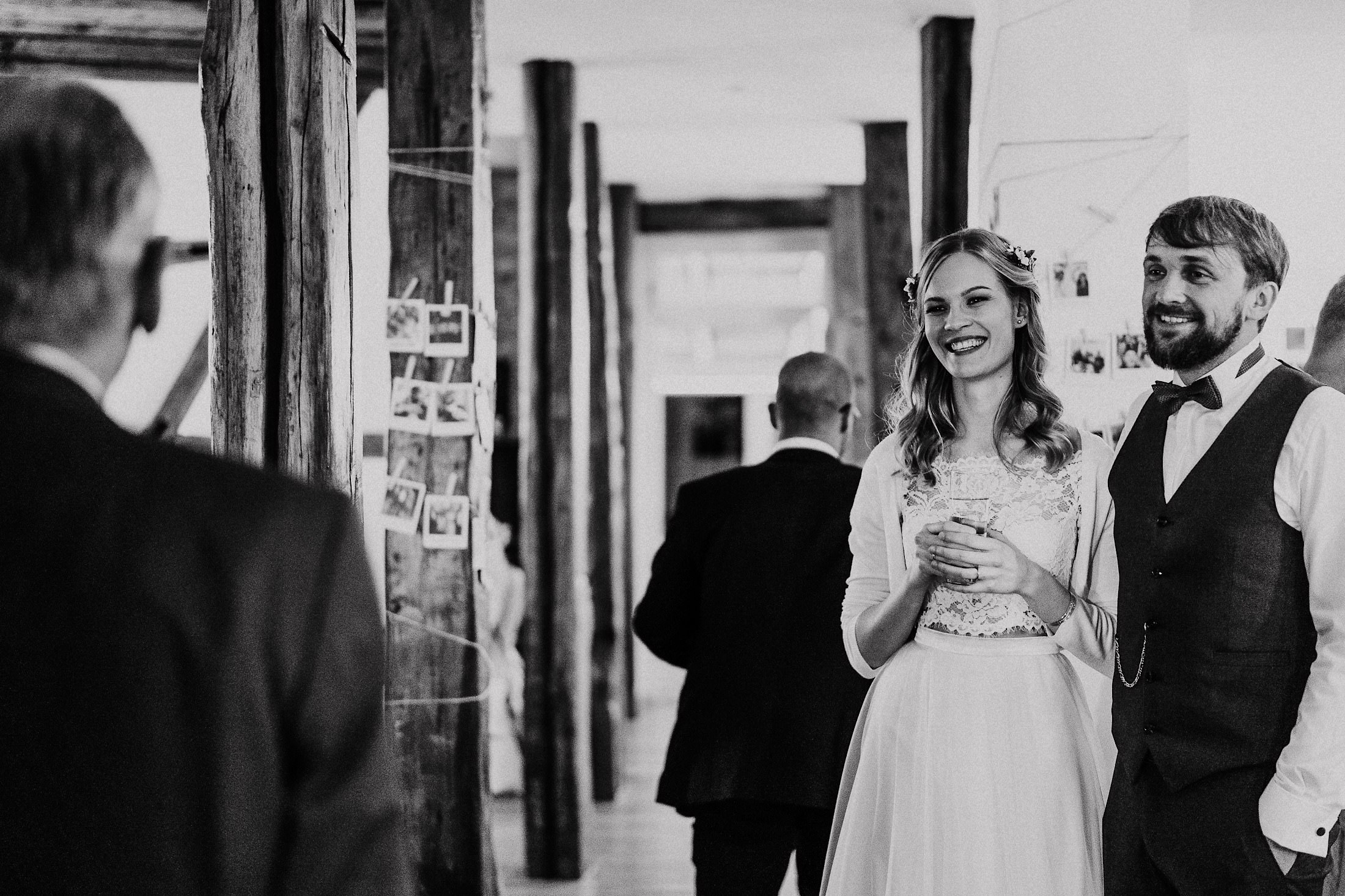 Braut und Bräutigam lachen in der Scheune der Eventmühle vor einer Wand mit Polaroids.