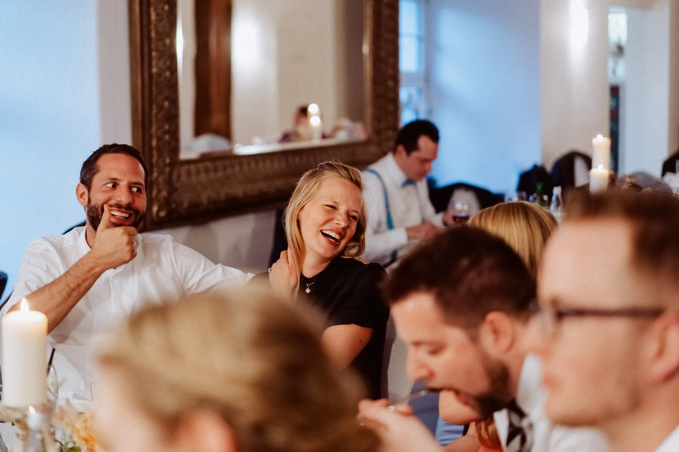 Hochzeitsgäste lachen an der Tafel im Festsaal, dahinter ein goldgerahmter Spiegel.