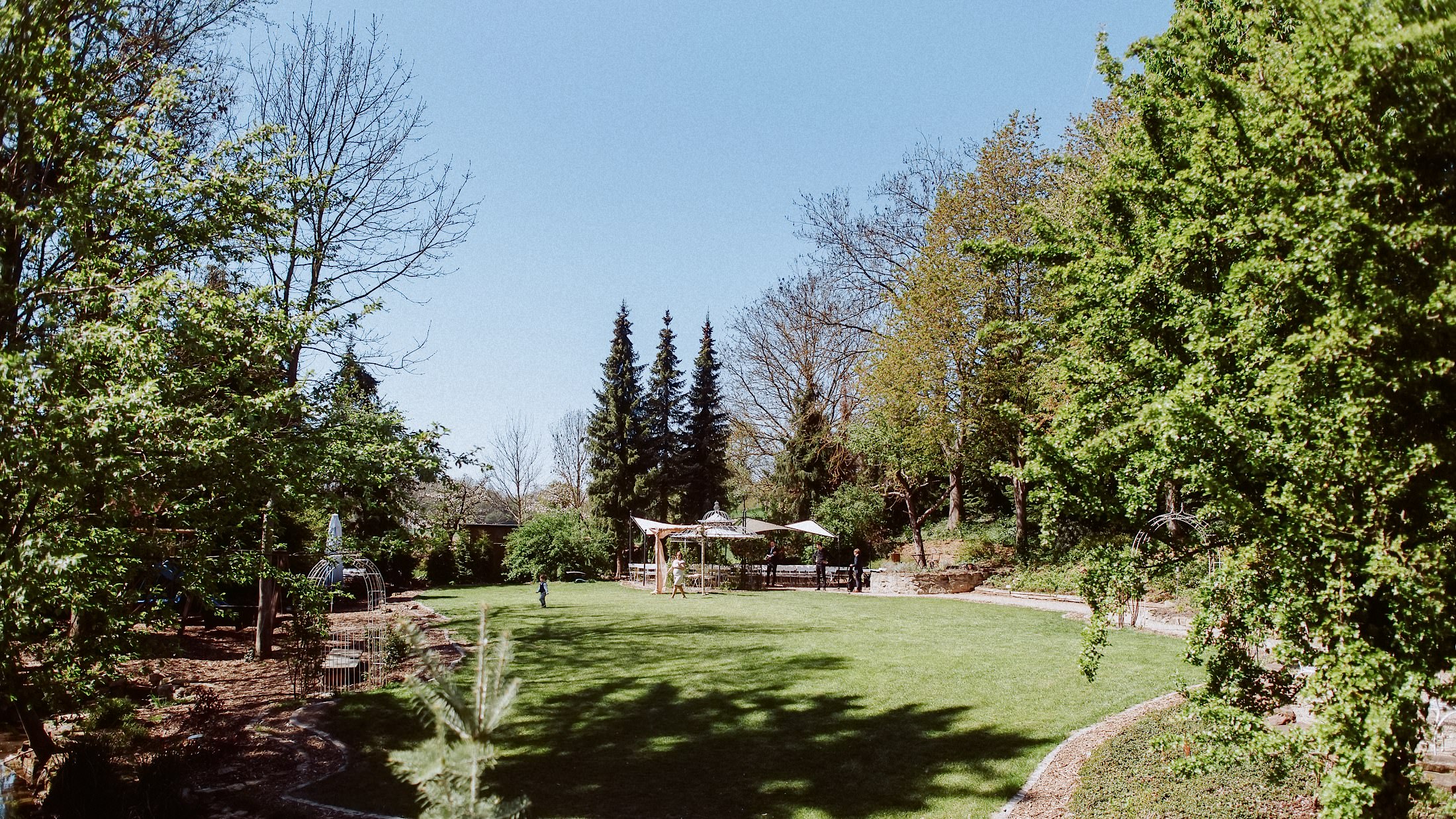 Gäste stehen unter der weißen Pergola im parkähnlichen Garten mit altem Baumbestand.