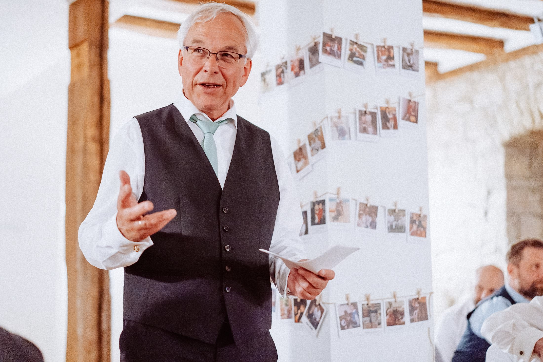 Ein älterer Gast hält eine Rede vor einer Wand mit angepinnten Fotos.