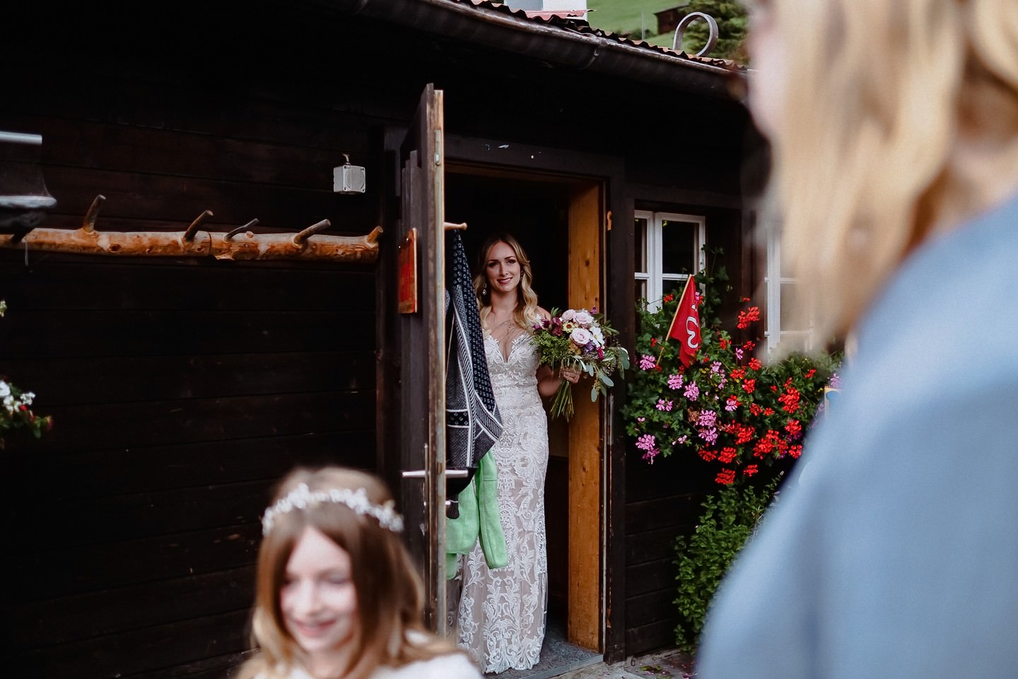 Braut im Spitzenkleid vor Holzchalet, Blumenstrauß, Geranien.