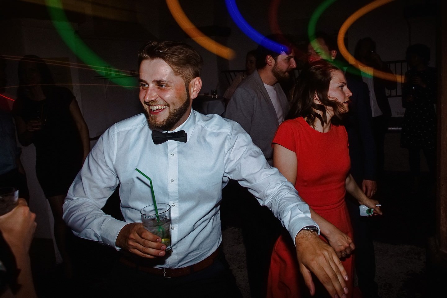 Bräutigam mit Drink und Gast in rotem Kleid lachen auf Tanzfläche, Laser-Lichter.