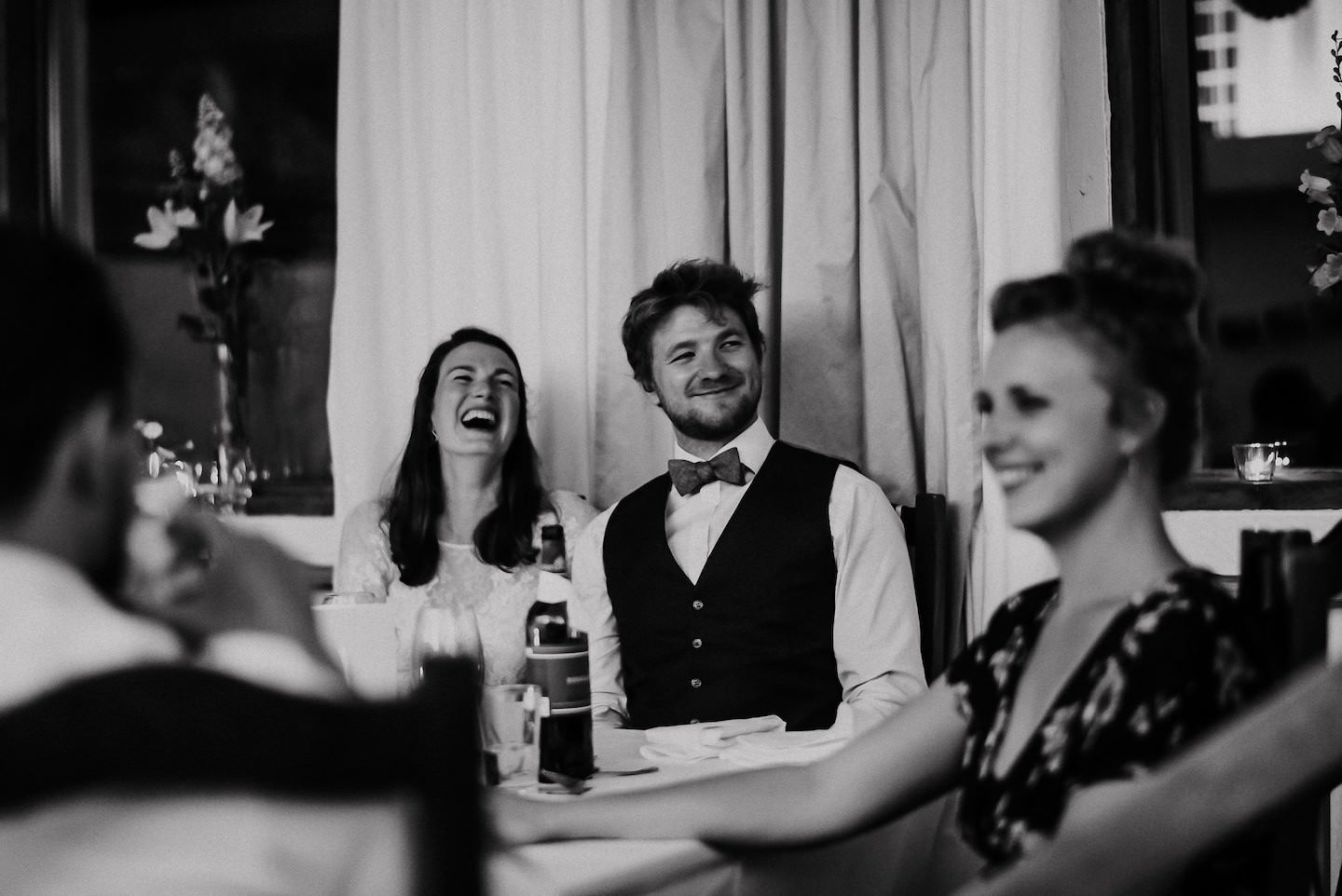 Brautpaar lacht zusammen bei Hochzeitstisch-Moment, schwarz-weiß, Gäste im Hintergrund.