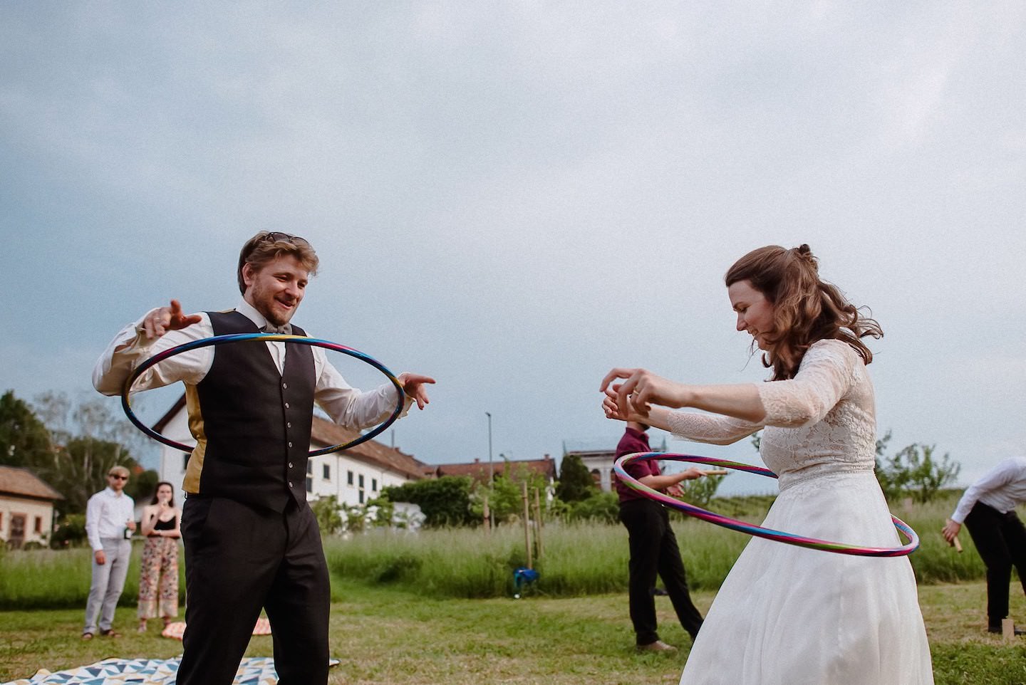 Brautpaar tanzt gemeinsam Hula-Hoop auf Wiese, fröhlich, Hochzeitsgäste beobachten.