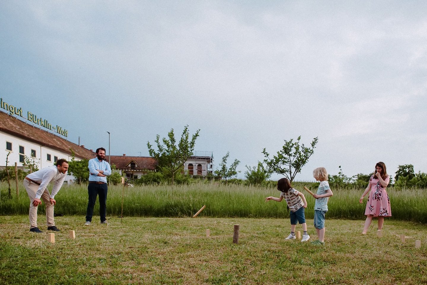 Gäste und Kinder spielen Holzklötze-Wurfspiel (Kubb) auf Hofgut-Rasen zusammen.