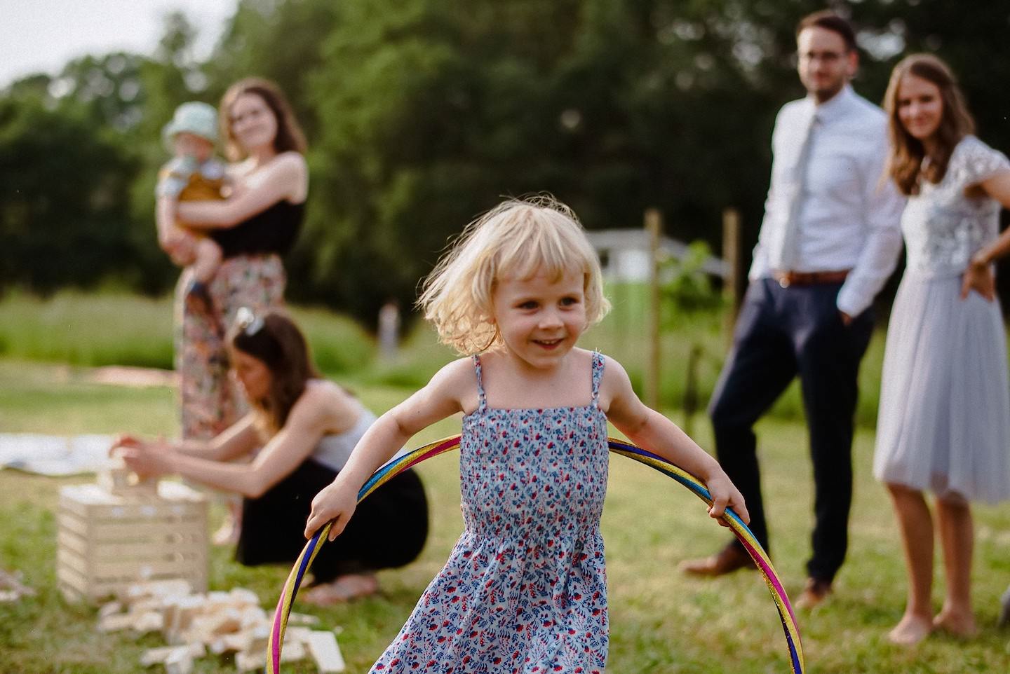 Kind mit Hula-Hoop auf Hofgut-Wiese tanzt, Hochzeitsgäste applaudieren, Sonne.