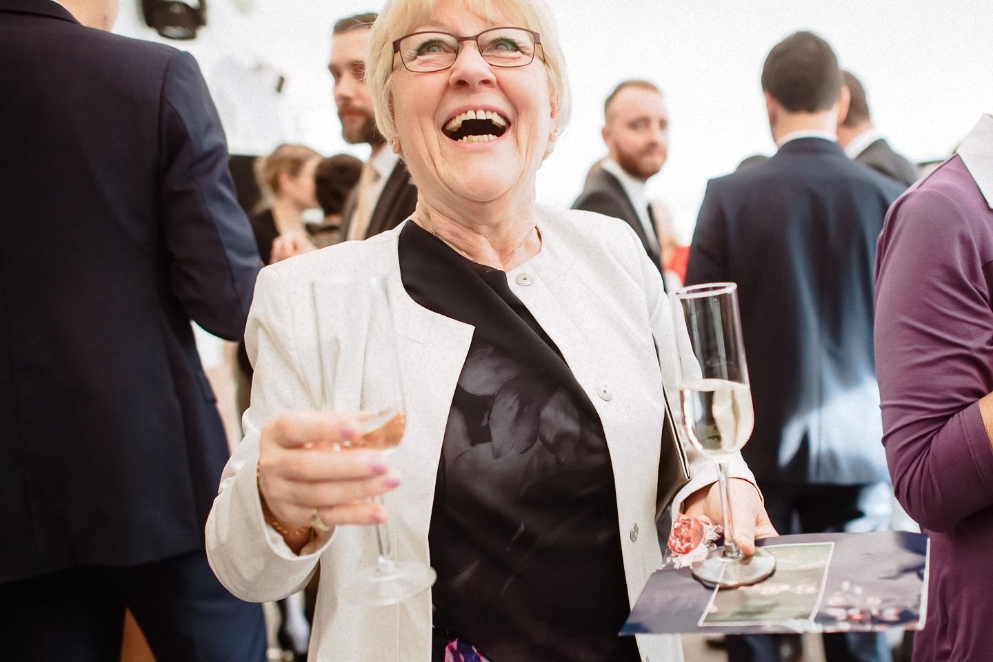 Ältere Frau mit Sekt-Glas, Gäste im Hintergrund feiern.