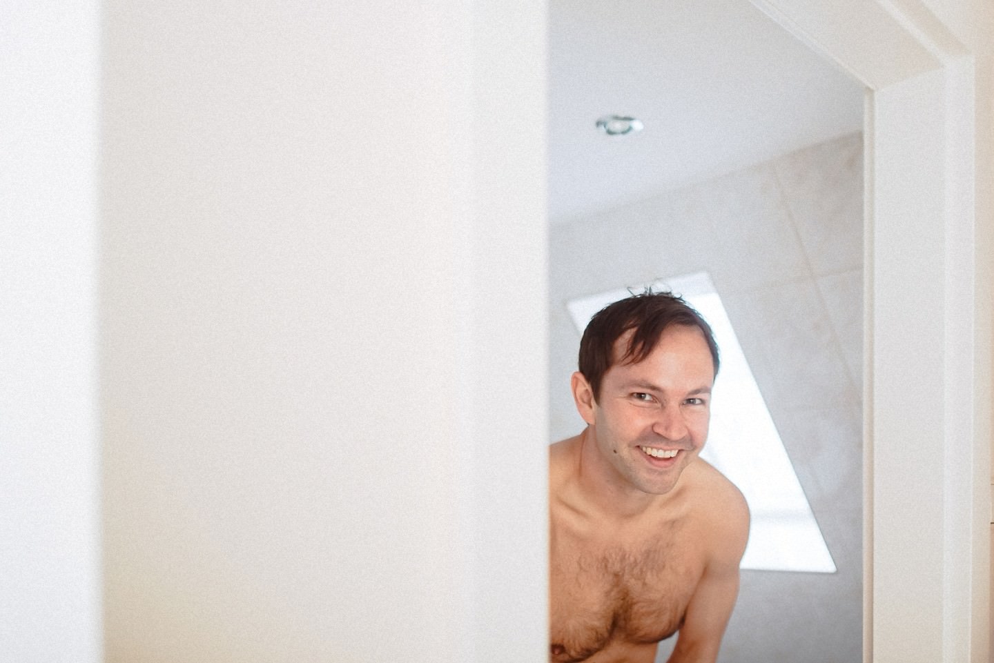 Bräutigam oben-ohne lächelt aus Bad/Dusche im hellen Raum.