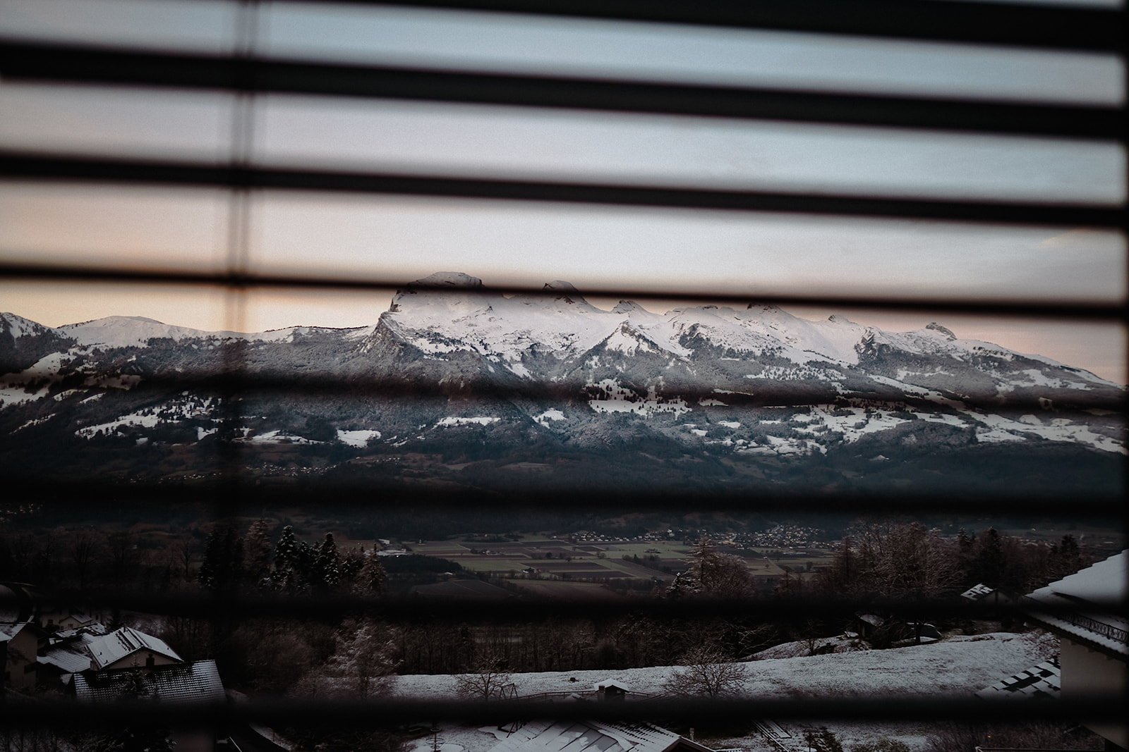 Blick durch die Jalousie auf die verschneiten Berge Liechtensteins im Morgenlicht