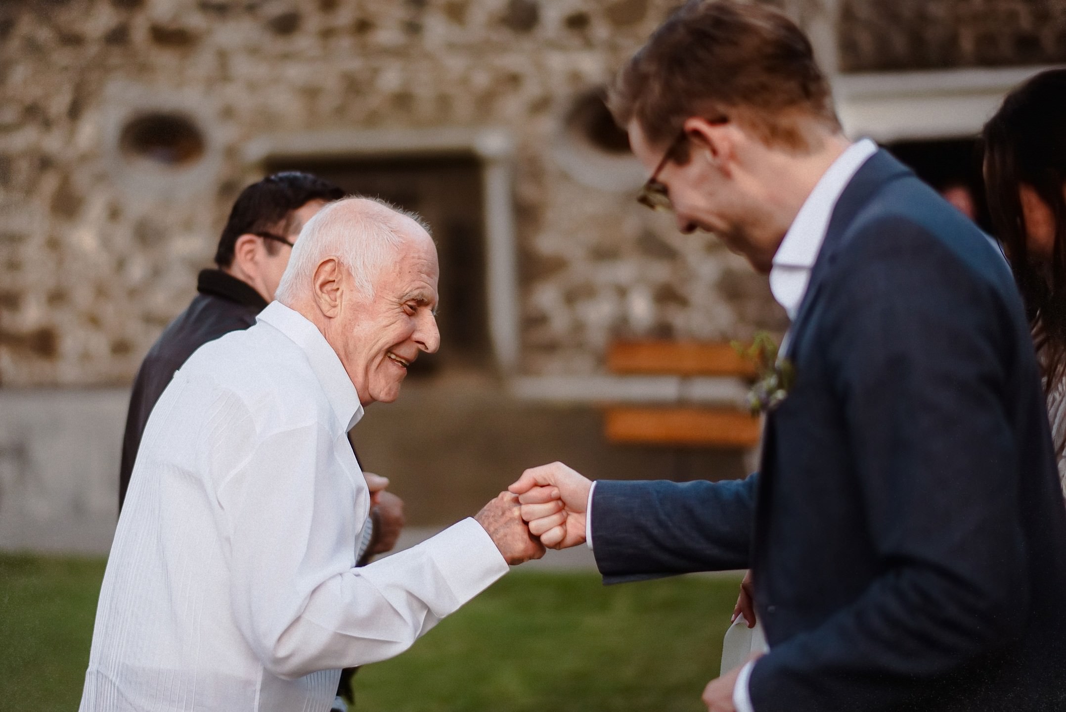 Ein älterer Mann schlägt fröhlich in die Hand des Bräutigams, Gäste schauen zu