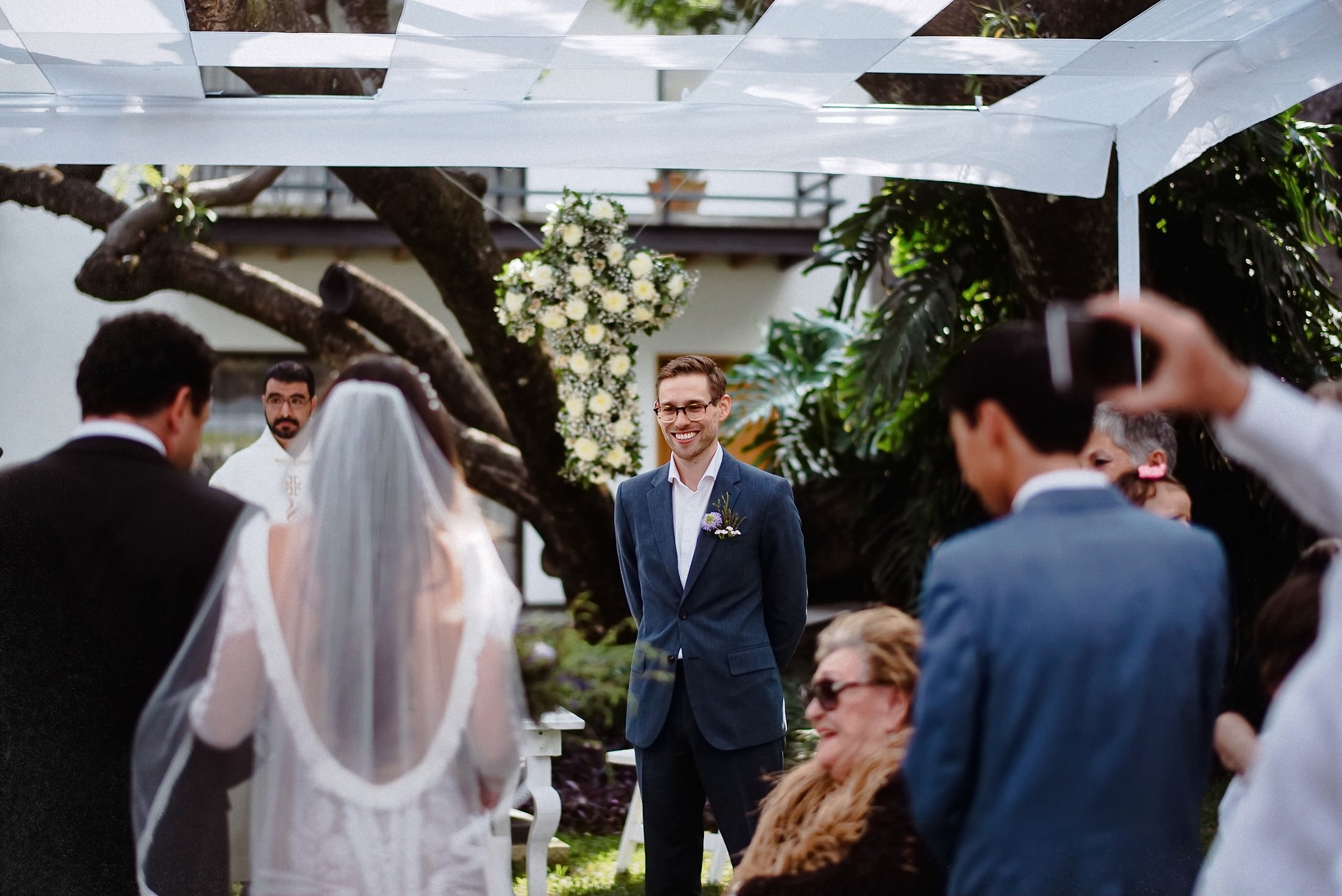 Der Bräutigam wartet beim First Look am Altar unter einer weißen Pergola auf die Braut mit Schleier