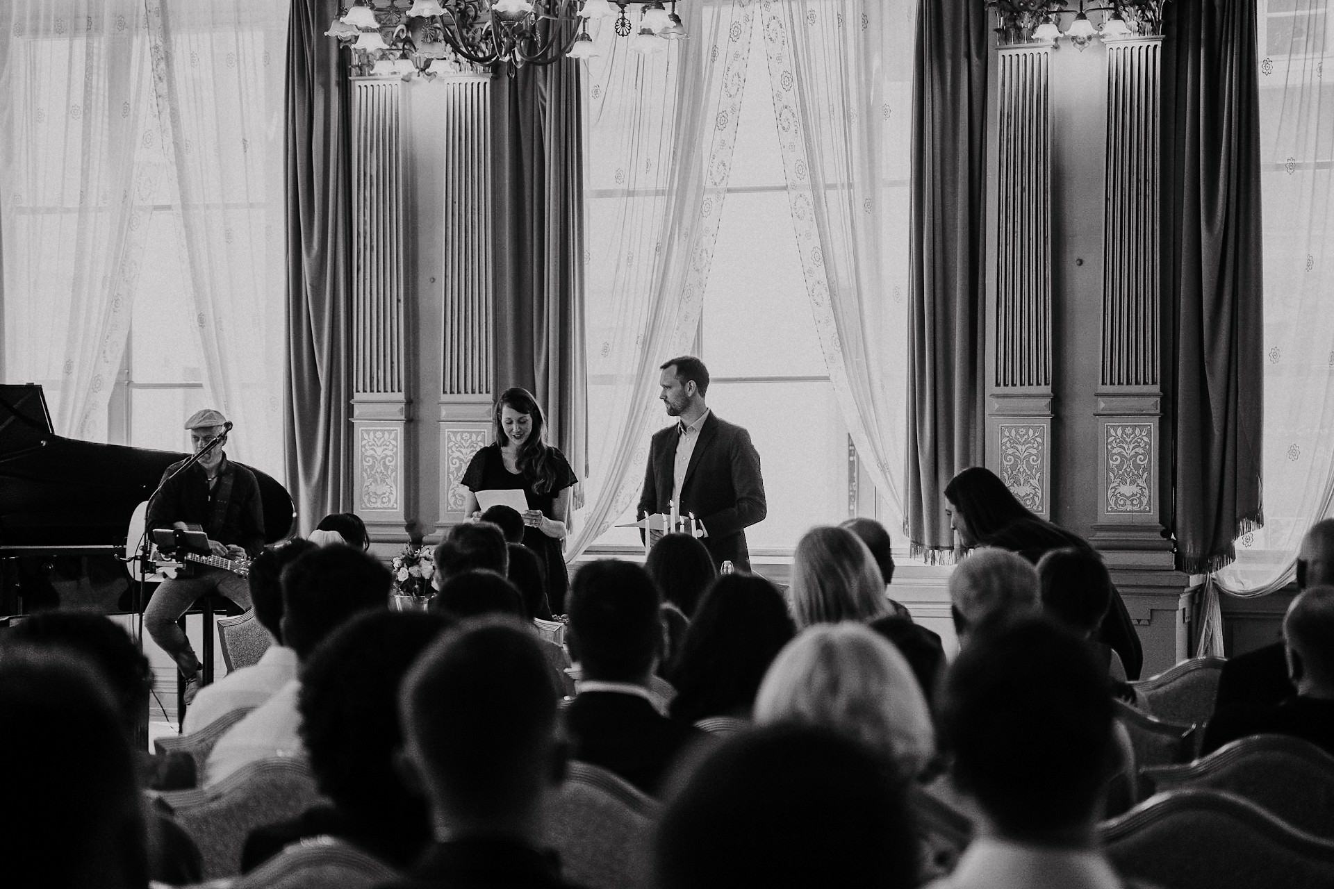 Freunde halten Rede auf Bühne im elegant dekorierten Trausaal mit Klavier.