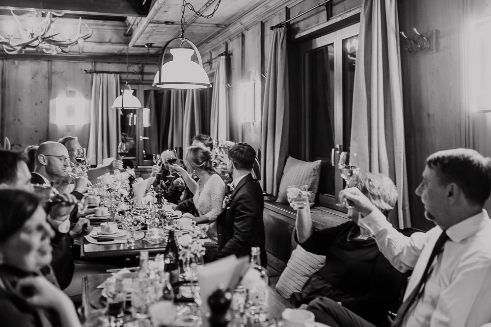 Die Hochzeitsgesellschaft hebt die Gläser an der langen Tafel in der Berghütte