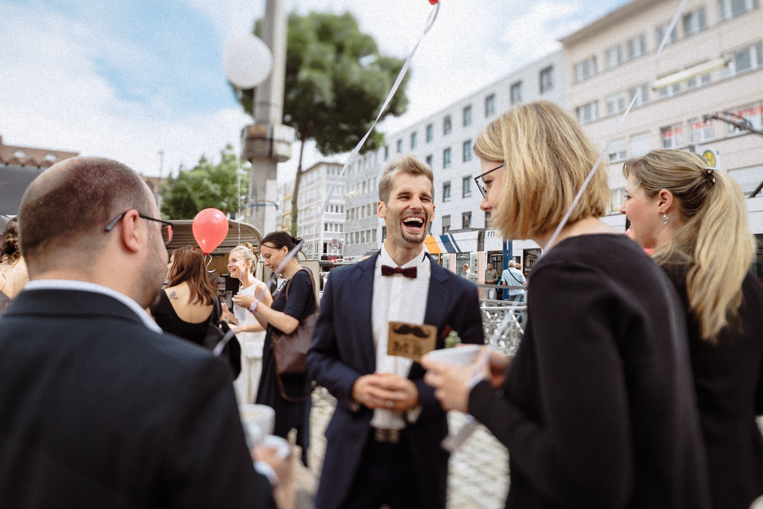 Bräutigam lacht mit Gästen beim Empfang unter freiem Himmel in der Innenstadt.