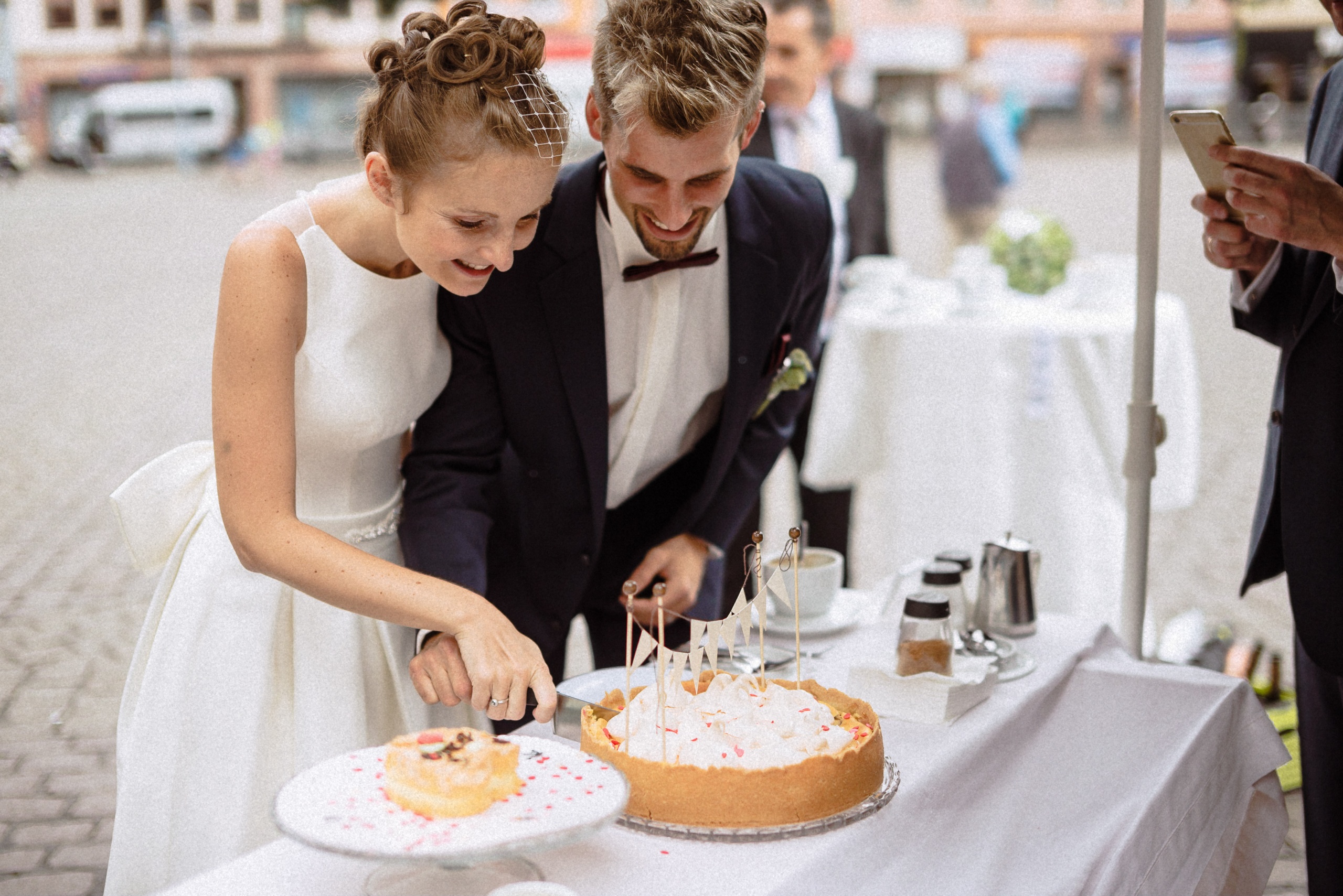 Brautpaar schneidet gemeinsam die Hochzeitstorte am Stehtisch auf dem Marktplatz an.