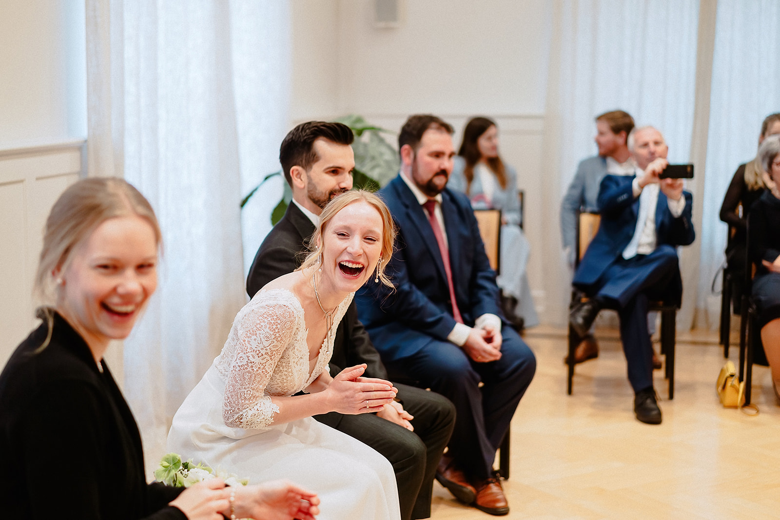 Braut und Bräutigam lachen im hellen Trauzimmer, dahinter applaudieren die Gäste