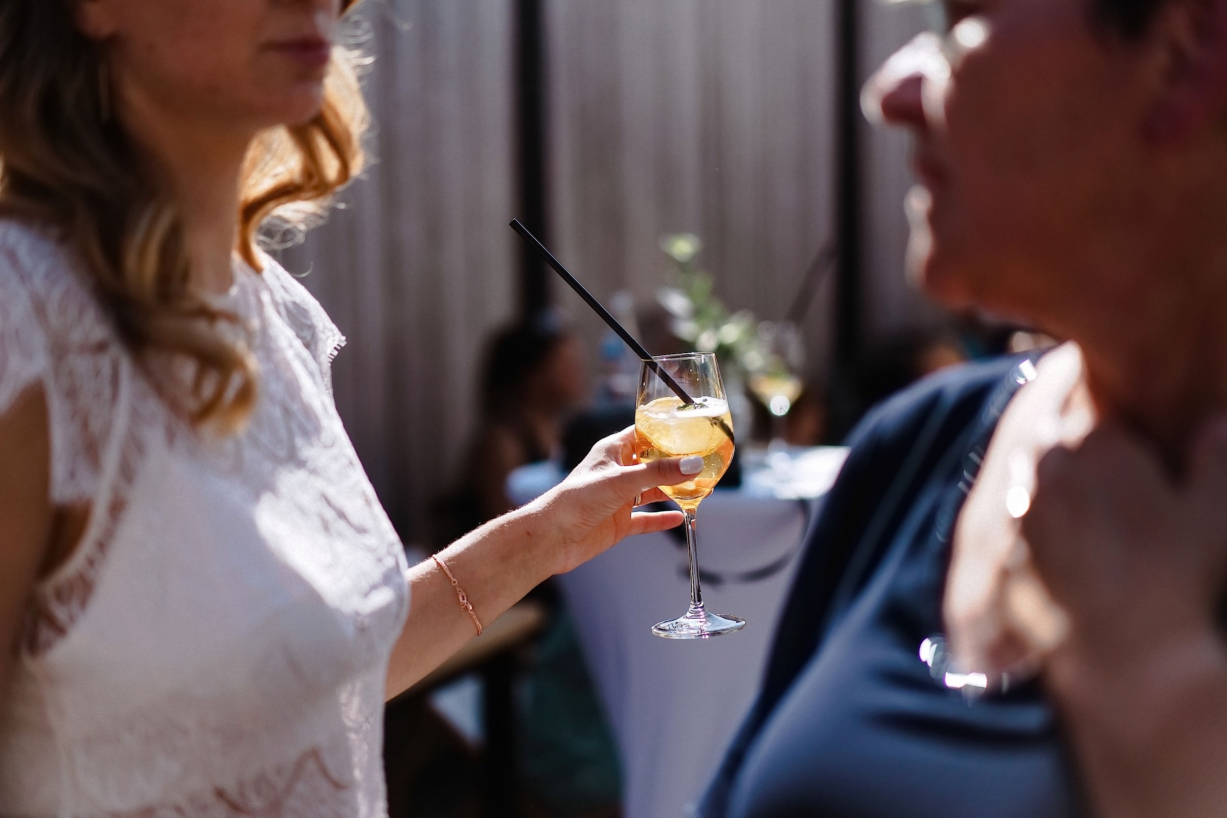 Die Braut hält einen Aperol Spritz in der Hand und unterhält sich im Gegenlicht mit einem Gast.