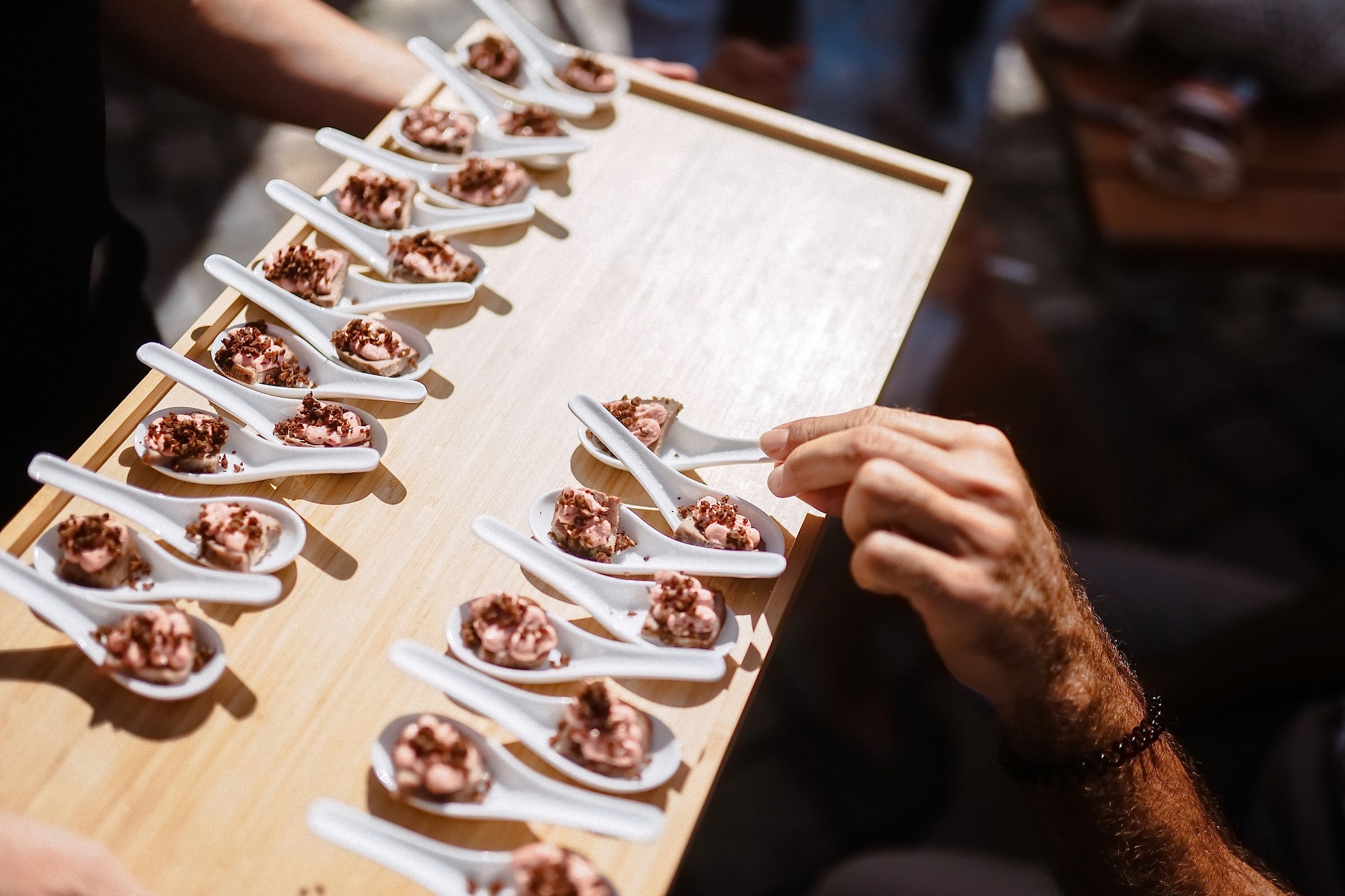 Ein Gast greift sich einen Fingerfood-Löffel mit Mousse und Krokant von einem servierten Holzbrett.