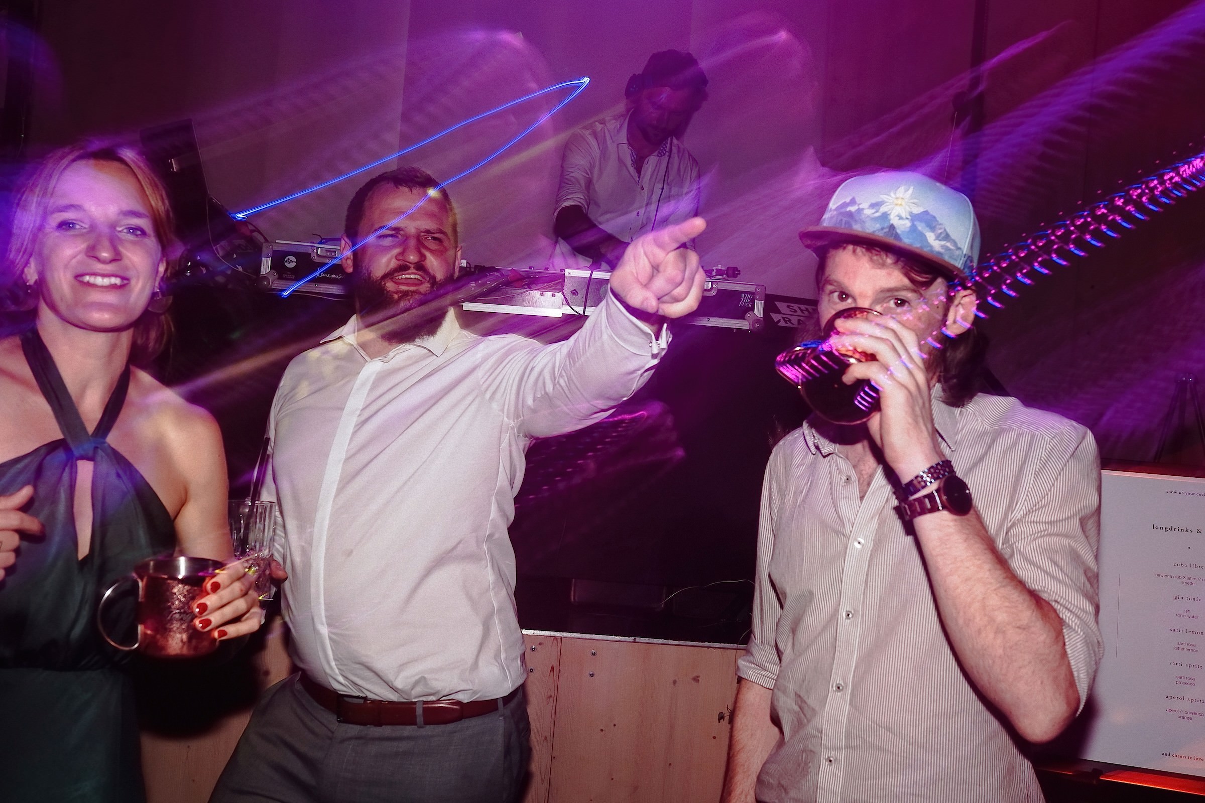 Gäste feiern ausgelassen an der DJ-Booth, während ein Mann in Trachtenkappe aus einem dunklen Glas trinkt.