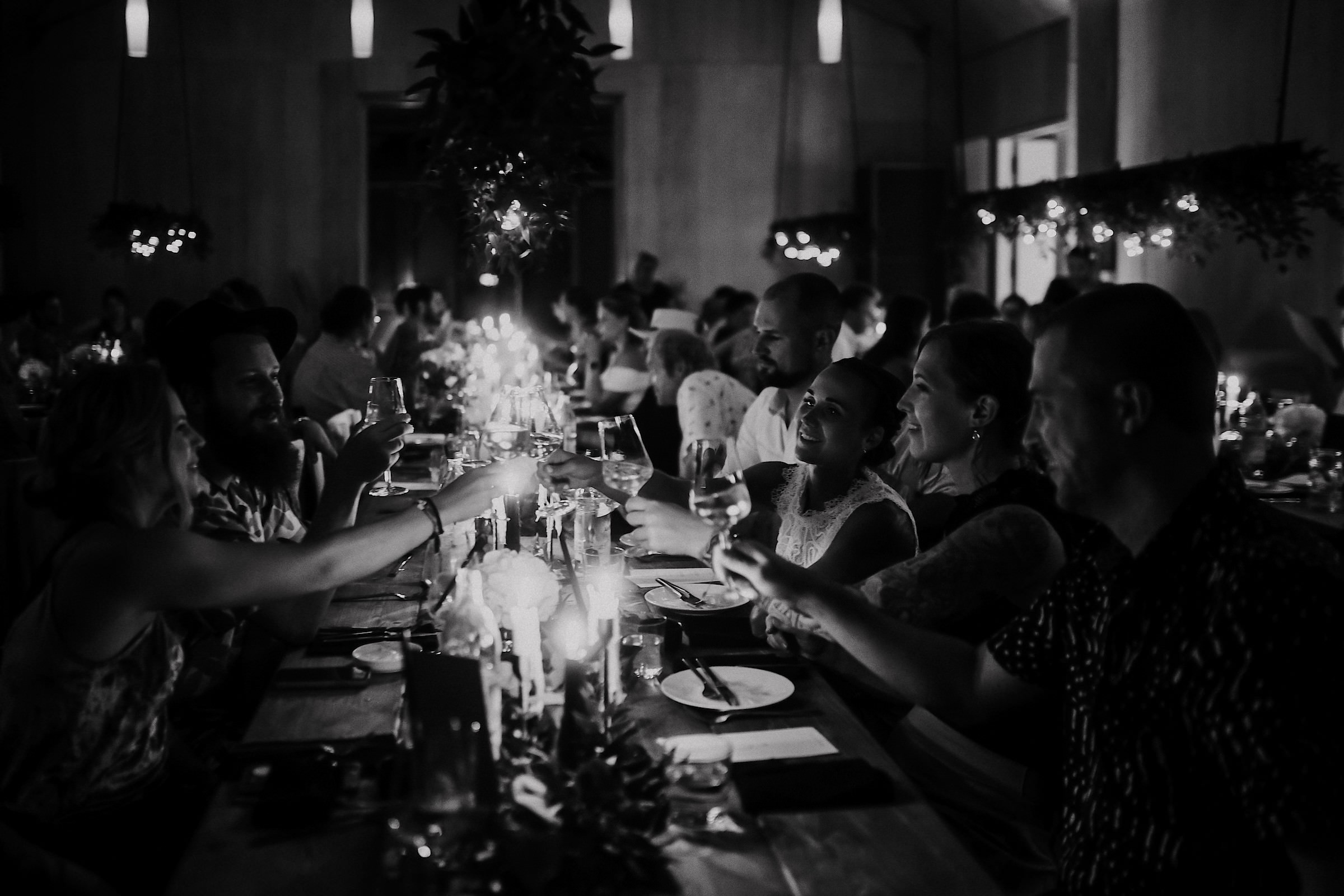 Schwarzweißaufnahme: Die Gästereihe an der langen Tafel stößt lachend mit Weingläsern an.