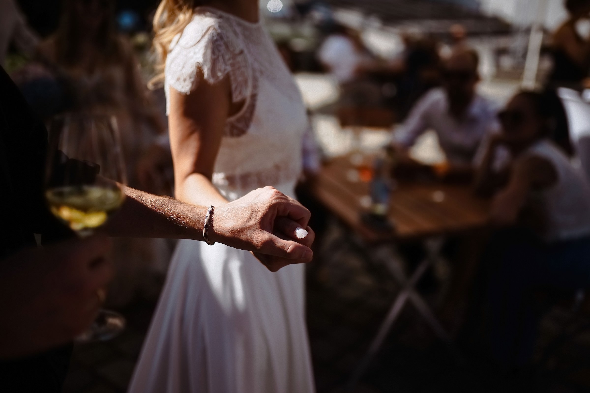 Der Bräutigam hält die Hand der Braut, während er ein Glas Weißwein in der anderen Hand hält und Gäste im Hintergrund an Tischen sitzen.