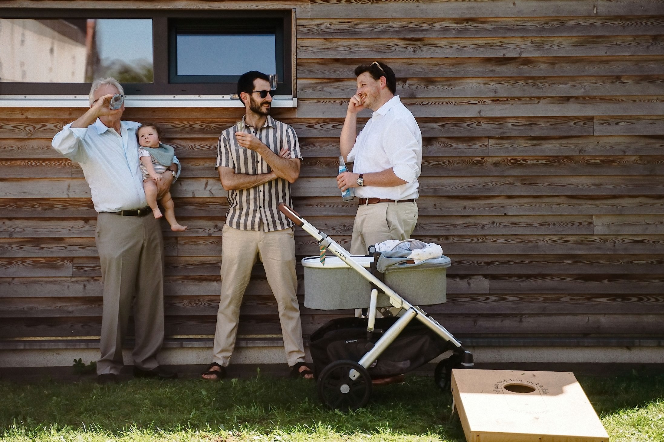 Drei Männer stehen mit Getränken an der Holzfassade, einer hält ein Baby im Arm, daneben ein Kinderwagen und ein Cornhole-Brett.