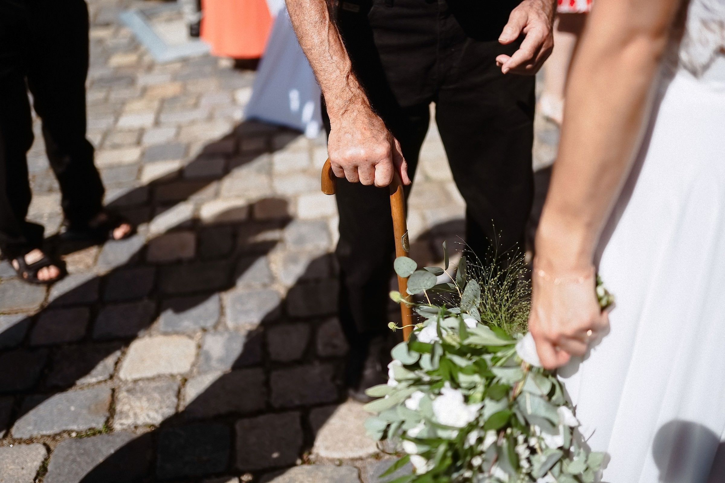 Detailaufnahme: Ein älterer Mann stützt sich auf seinen Gehstock neben der Braut, die einen Blumenstrauß aus Eukalyptus und weißen Blüten trägt.