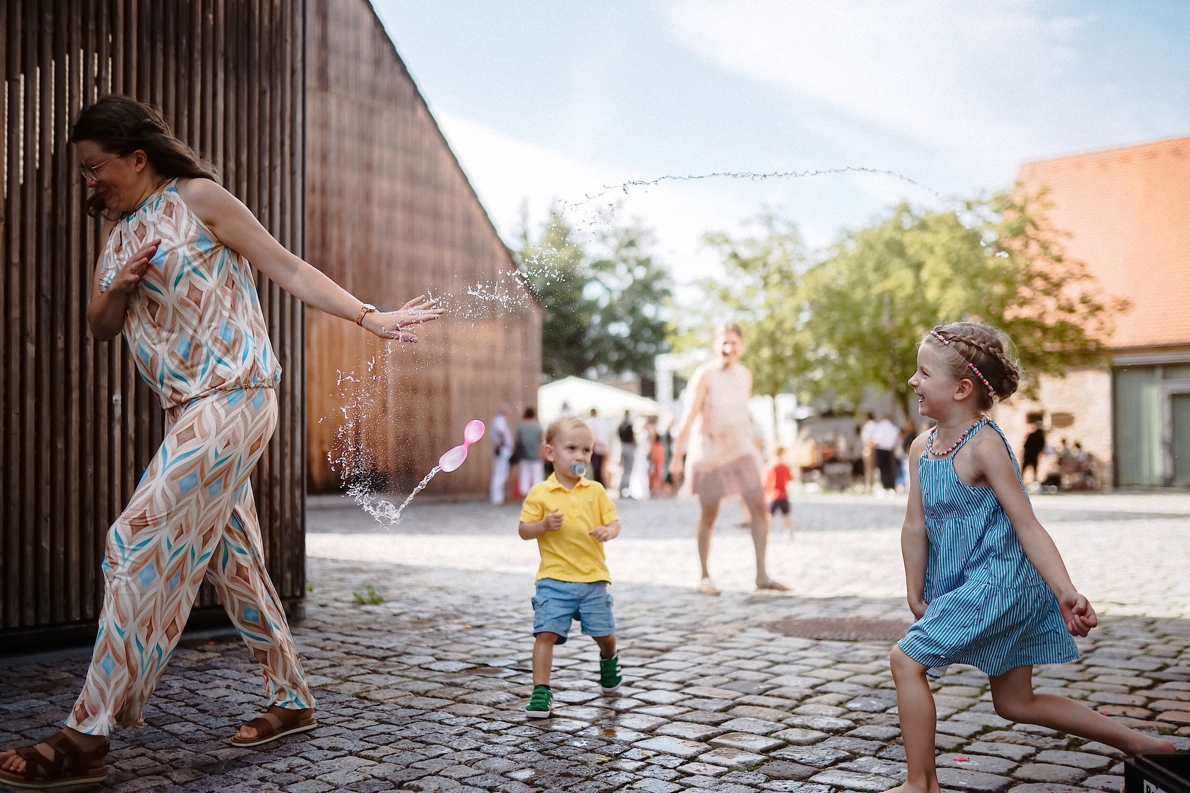 Eine Frau weicht lachend einem Wasserstrahl aus, während Kinder auf dem Kopfsteinpflaster spielen.