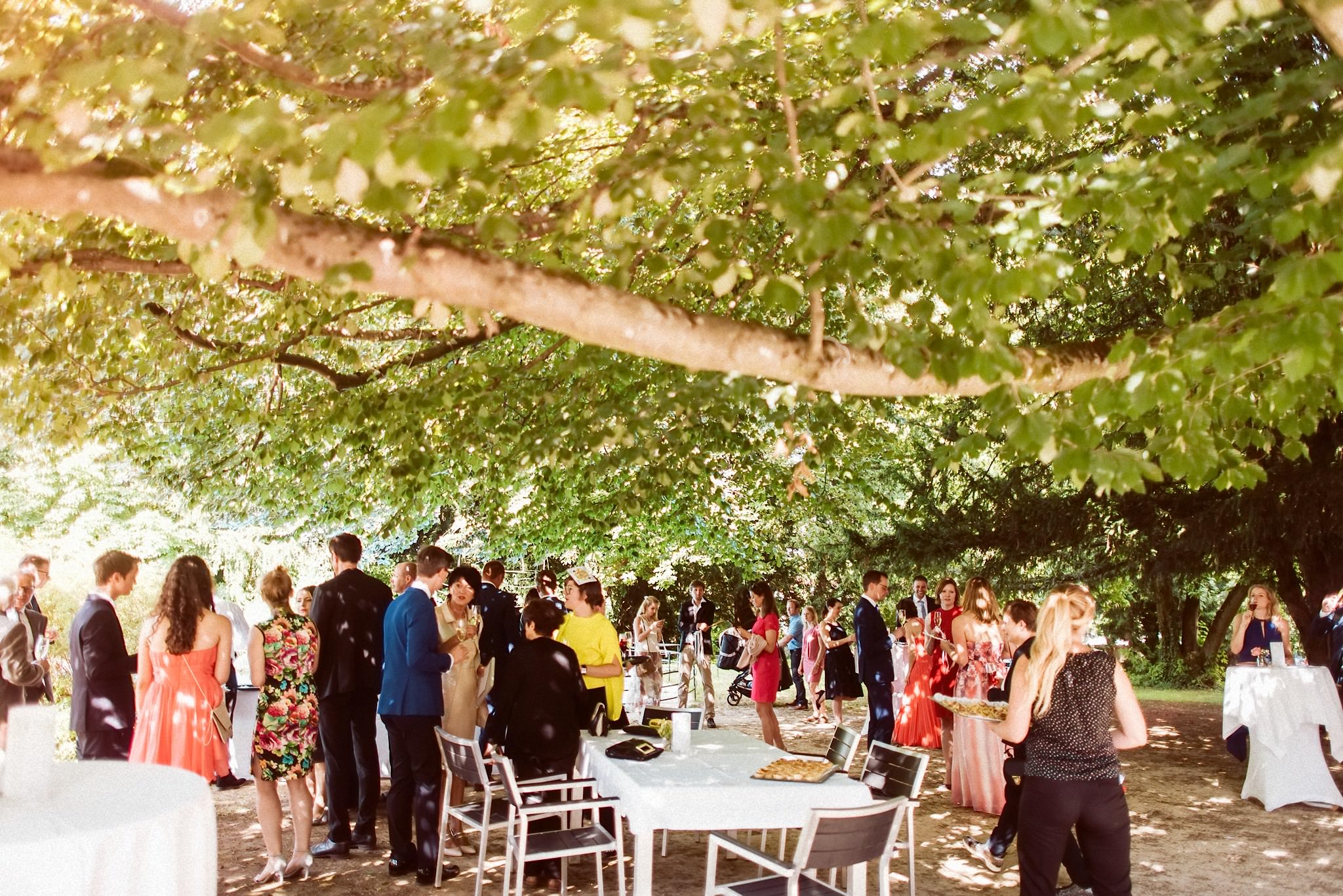 Die Hochzeitsgesellschaft feiert den Sektempfang unter Bäumen im Schlosspark.
