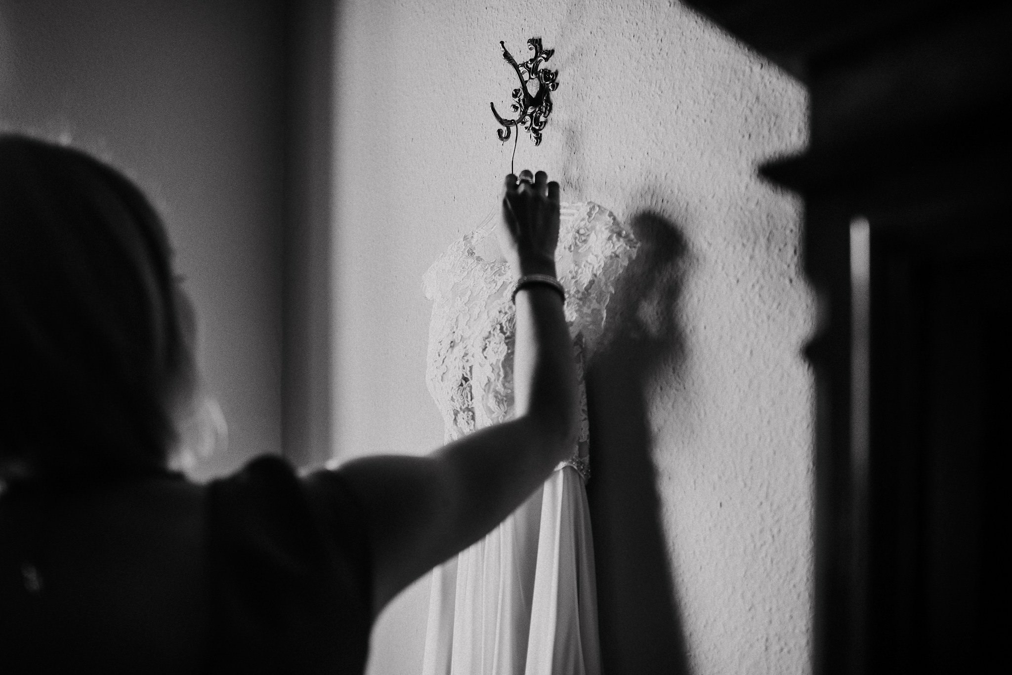 Eine Frau hängt das Spitzen-Brautkleid in dramatischem Licht an einen schmiedeeisernen Wandhaken.