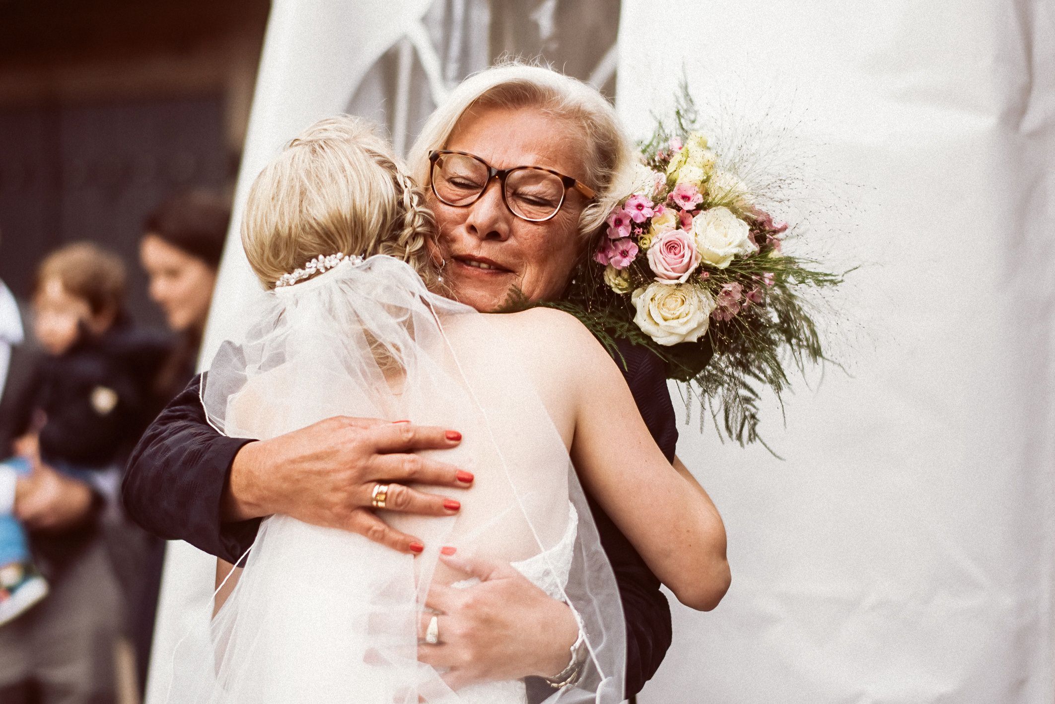 Ältere Frau mit Brille umarmt Braut herzlich mit Blumen und Freude.