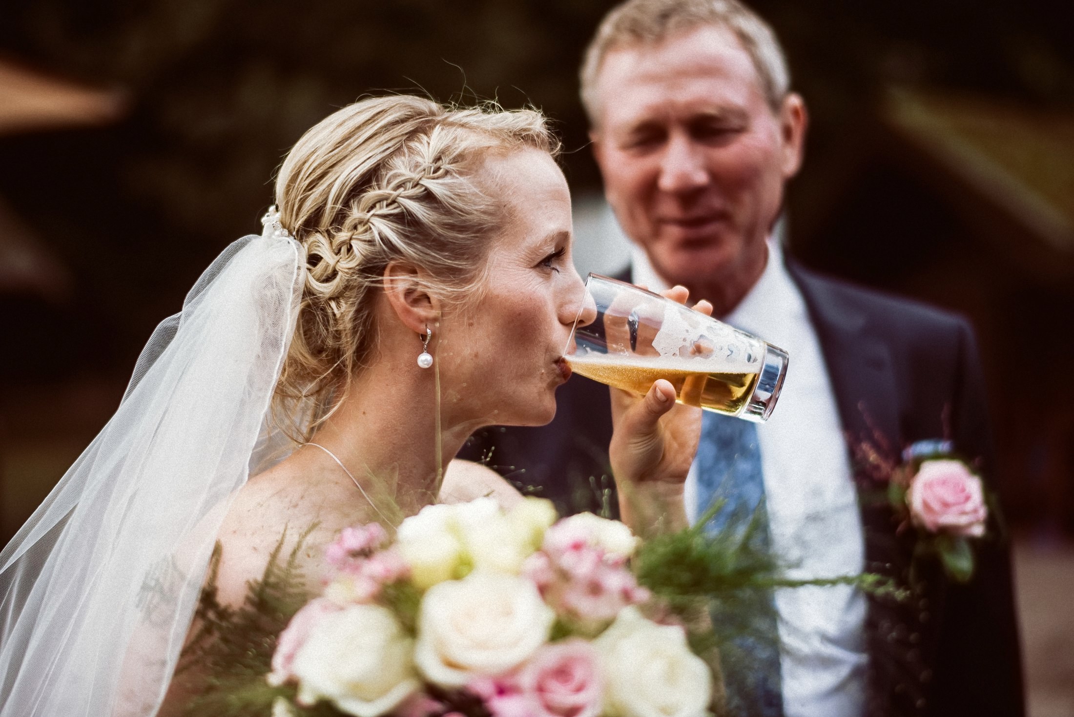 Die Braut trinkt genüsslich ein Bier, ein älterer Gast schaut amüsiert zu.