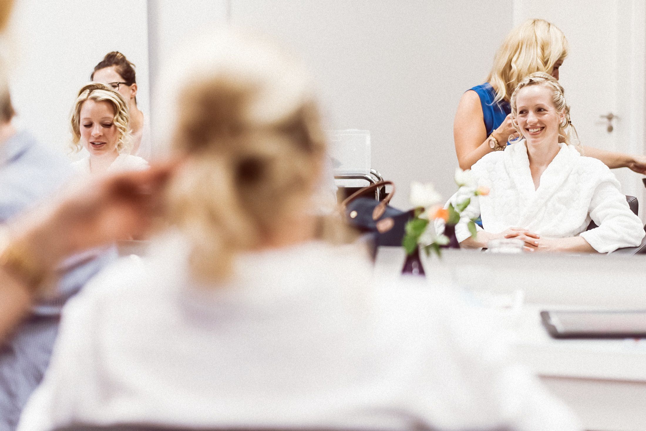 Braut und Helferinnen lachen bei Frisur- und Make-up-Vorbereitung im hellen Zimmer.