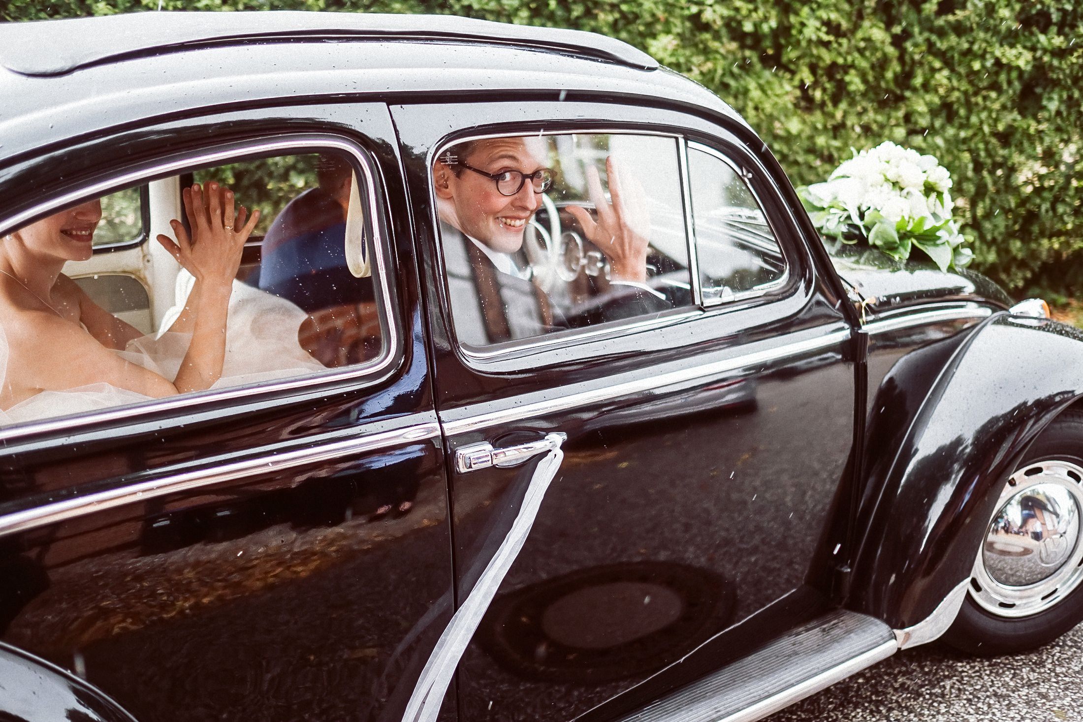 Braut winkt freudig aus schwarzem VW Käfer, Bräutigam lenkt das Hochzeitsauto.
