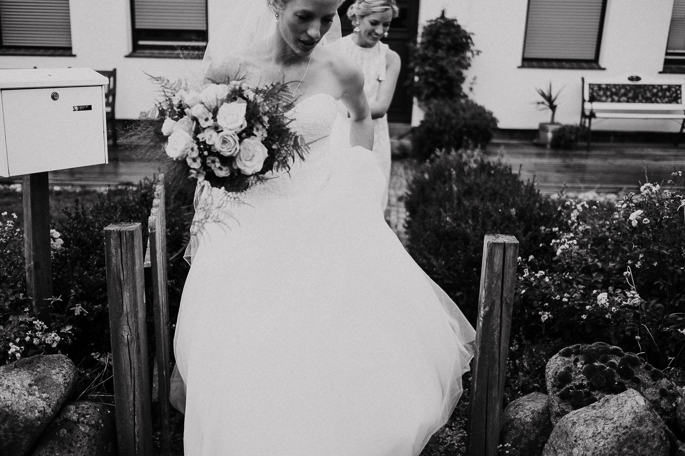 Braut von hinten im weißen Kleid mit üppigem Brautstrauß vor weißem Haus.