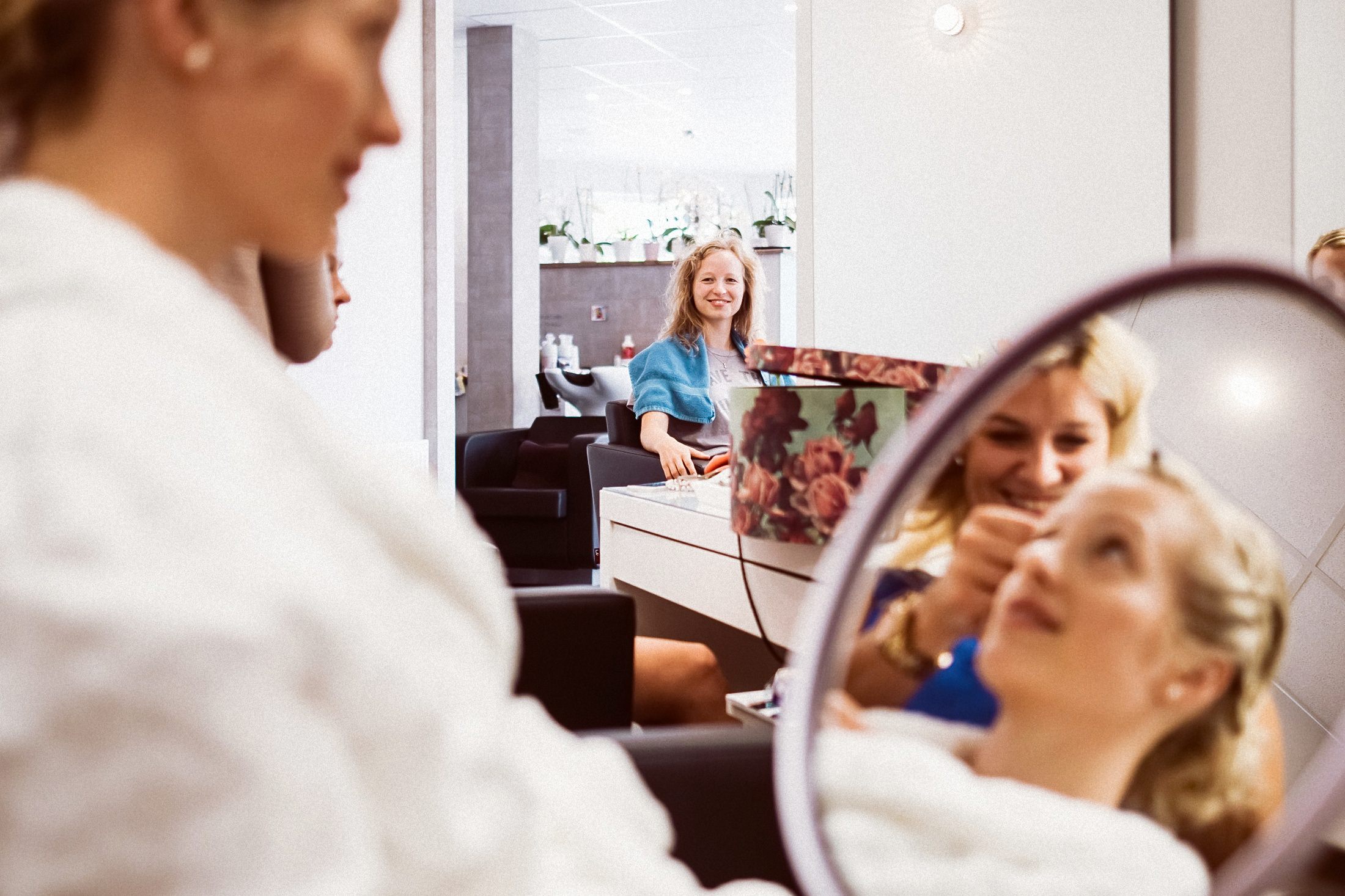 Frau schaut freudig in Spiegel während Freundin im hellen Kosmetik-Zimmer neben sitzt.
