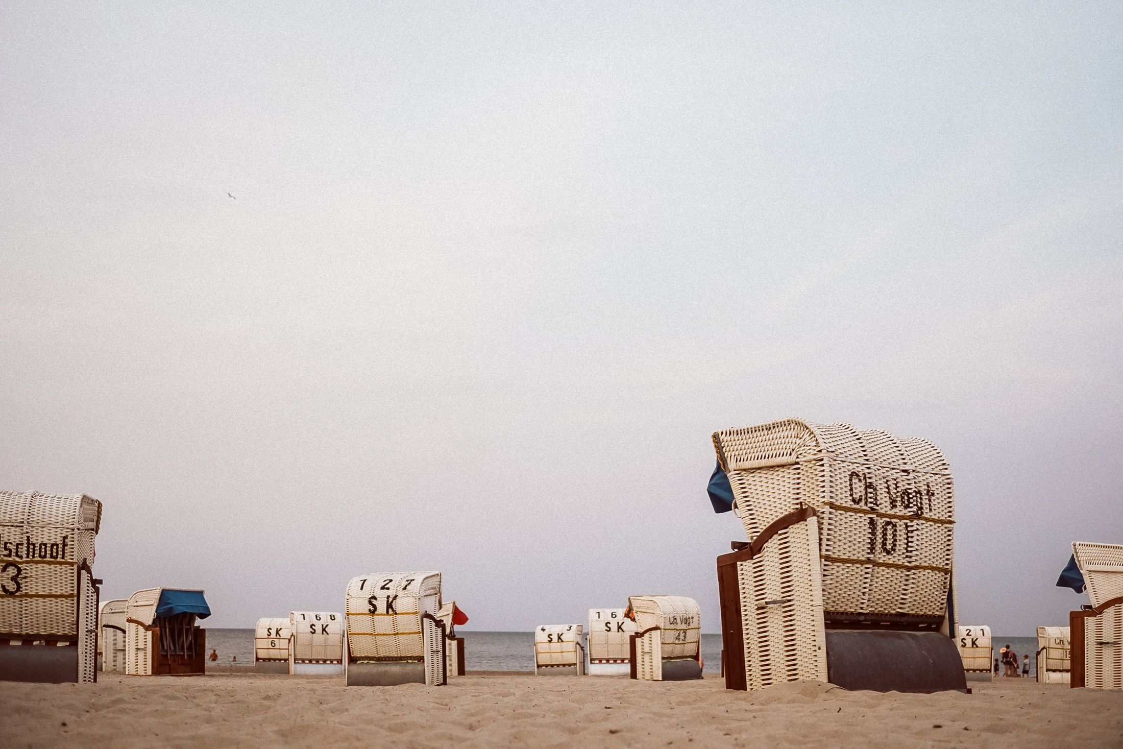 Reihe heller Strandkörbe am Ostseestrand im Sand unter bedecktem Himmel.
