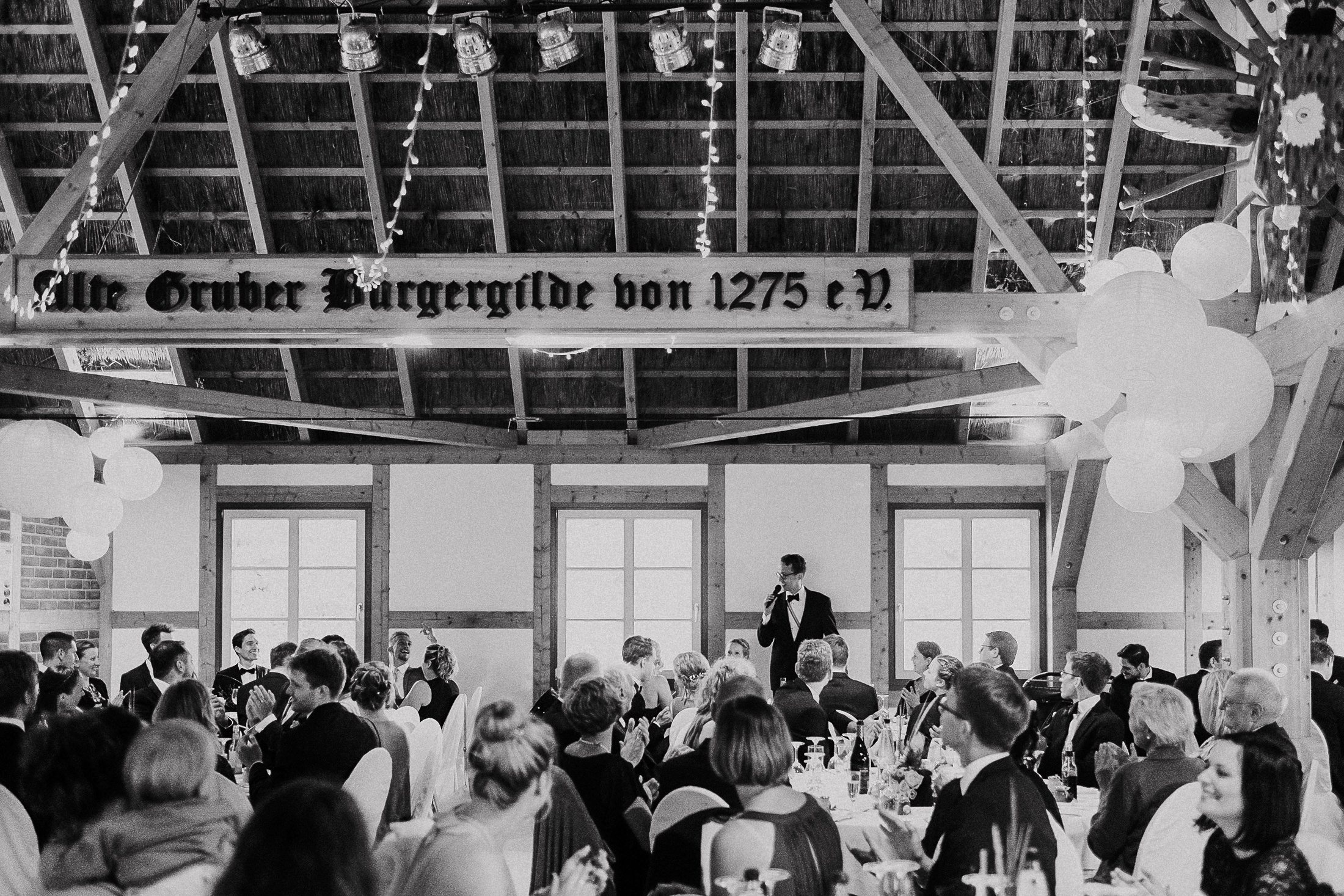 Schwarz-Weiß: Bräutigam hält Rede vor vollbesetztem Festzelt mit Balken.
